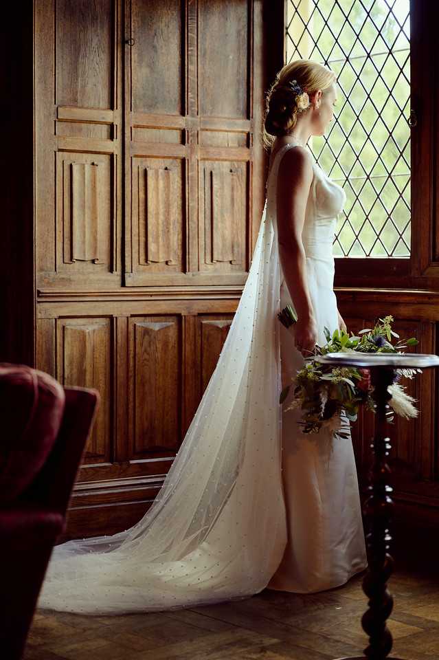 A bridal portrait taken indoors in a room featuring dark oak wood paneling and a leaded diamond-pane window. The bride stands in profile, looking out the window, wearing a fitted ivory sleeveless gown with a long cathedral-length veil dotted with small embellishments. Her hair is styled in a low updo with small floral hair accessories. She holds a loosely arranged bouquet featuring dark foliage, thistles, greenery, and pampas grass in muted, earthy tones. A dark wood pedestal table and a burgundy upholstered chair are visible in the frame. The room's warm wood tones and period furnishings suggest a historic chateau or manor house interior. The shot is a full-length portrait with natural window light illuminating the bride from the side.
