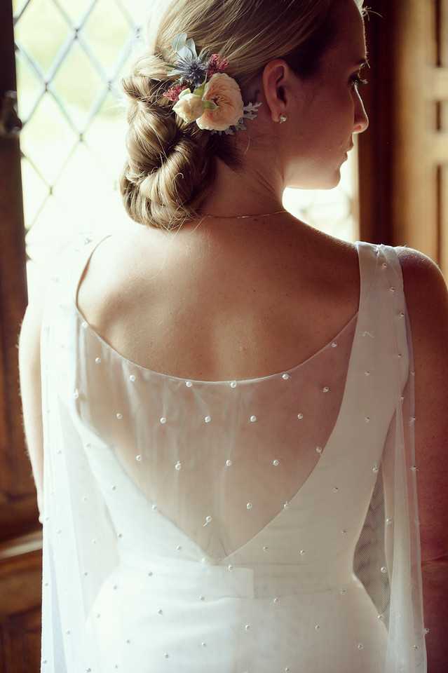 A close-up portrait of a bride shot from behind, highlighting the back detail of her white wedding dress, which features a sheer tulle overlay scattered with small pearl embellishments and a low scoop back. Her hair is styled in a twisted low chignon adorned with a small floral hair piece containing blush ranunculus, dusty blue thistles, and deep pink accent blooms. She wears small stud earrings and a delicate necklace. The setting appears to be indoors near a leaded glass window that provides warm, diffused natural light, suggesting a historic building such as a chateau or manor house.