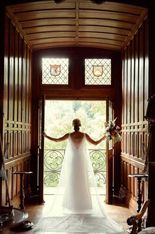 A bride stands with her back to the camera in a doorway of a richly wood-paneled interior room, arms outstretched to hold open two large wooden doors that open onto a wrought-iron balcony railing with garden views beyond. She wears a white A-line gown with a delicate dotted tulle train and a low back with sheer detailing, and her hair is styled in an updo with floral accessories. In her right hand she holds a loosely arranged bouquet featuring dried and fresh blooms in terracotta, ivory, and muted peach tones. Above the doorway are two stained-glass heraldic windows featuring coat-of-arms motifs in red, blue, and amber, set within dark wood framing with a barrel-vaulted ceiling — details consistent with a historic French chateau interior. The shot is a full-length portrait taken from inside the room, backlit by natural light flooding through the open doors, creating a strong silhouette contrast against the dark wood surroundings. Potential venue feature image.
