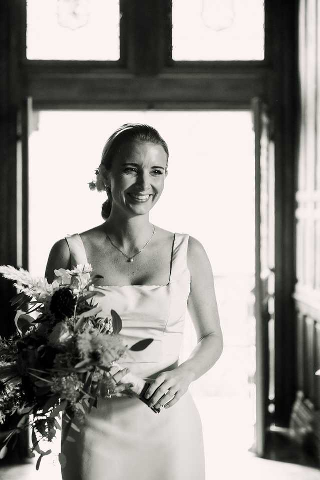 This is a black-and-white bridal portrait shot indoors, with the bride backlit by a large doorway or window that creates strong contrast between bright highlights and deep shadows. The bride wears a sleek, minimalist slip-style dress with wide shoulder straps and a square neckline, paired with a delicate pendant necklace and a small floral hair accessory. She holds a full, loosely arranged bouquet featuring large blooms, pampas grass-like textures, and foliage, displayed with high contrast in the monochrome tones. The composition is a close-up portrait framing her from approximately the waist up, with ornate dark wooden architectural framing visible in the background.
