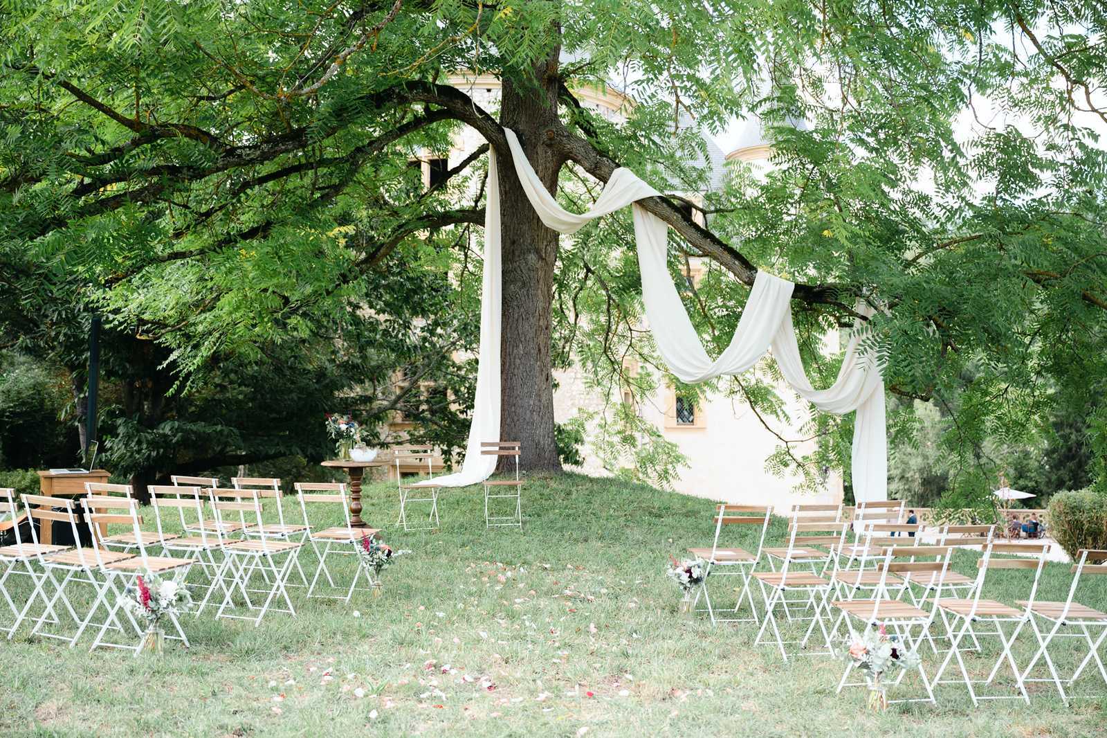 An outdoor wedding ceremony setup photographed before guests arrive, set on a lawn in the grounds of what appears to be a French château or manor house visible in the background. The ceremony backdrop is formed by a large mature tree with ivory or white sheer fabric draped across its branches, creating a natural altar effect accented with a few tropical-style leaves. Rows of white-framed wooden folding chairs are arranged on either side of a central aisle, with small mixed floral arrangements in pink, red, and white tones placed at the end of select rows. A small wooden pedestal table with a floral arrangement and what appears to be a ceremony book sits to the left of the tree, and scattered flower petals line the aisle. The overall styling is romantic-natural with a light, airy palette of white and wood tones. Wide shot capturing the full ceremony layout. Potential venue feature image.