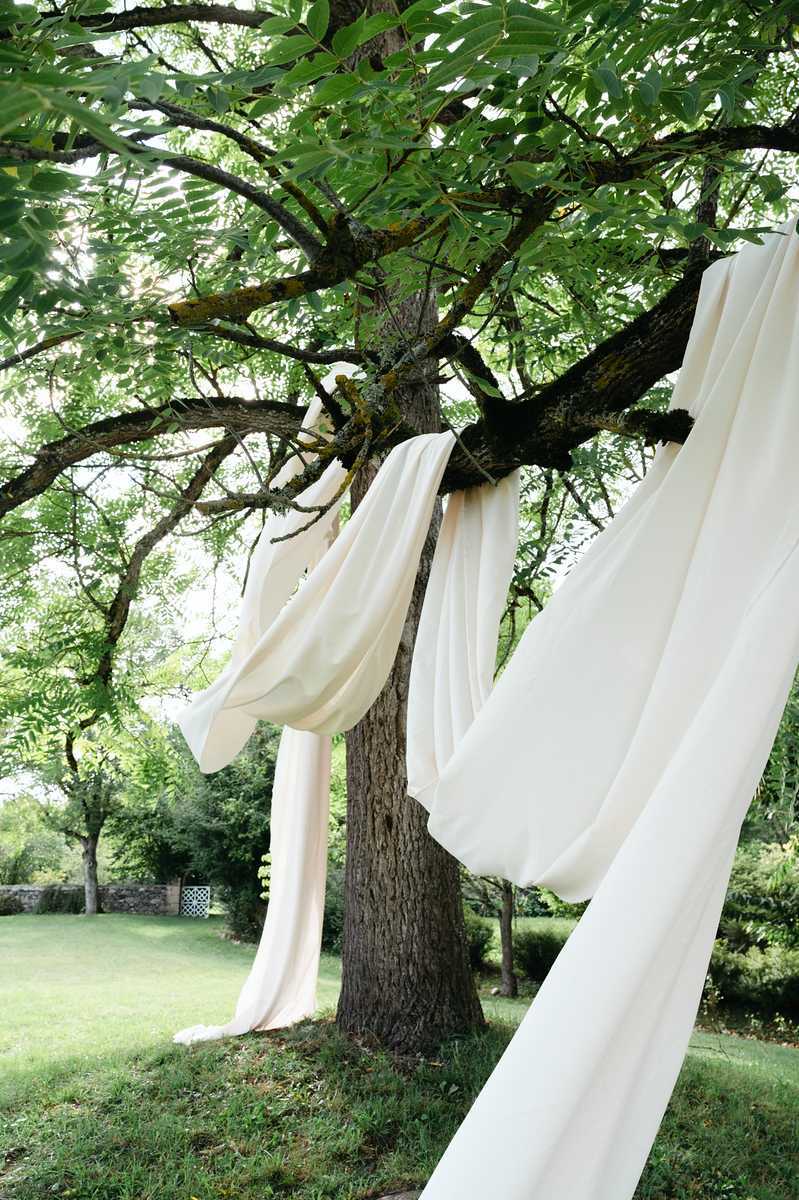 Outdoor ceremony or event space detail shot showing ivory/cream fabric draping draped over and hanging from the branches of a large mature tree in a garden setting. The flowing fabric panels are gathered and looped across multiple branches, cascading down toward the ground in soft folds. The setting appears to be a landscaped garden with a low stone wall visible in the background, consistent with a French country estate or chateau grounds. The composition is a medium-wide detail shot focused on the decorative fabric installation with no people visible.