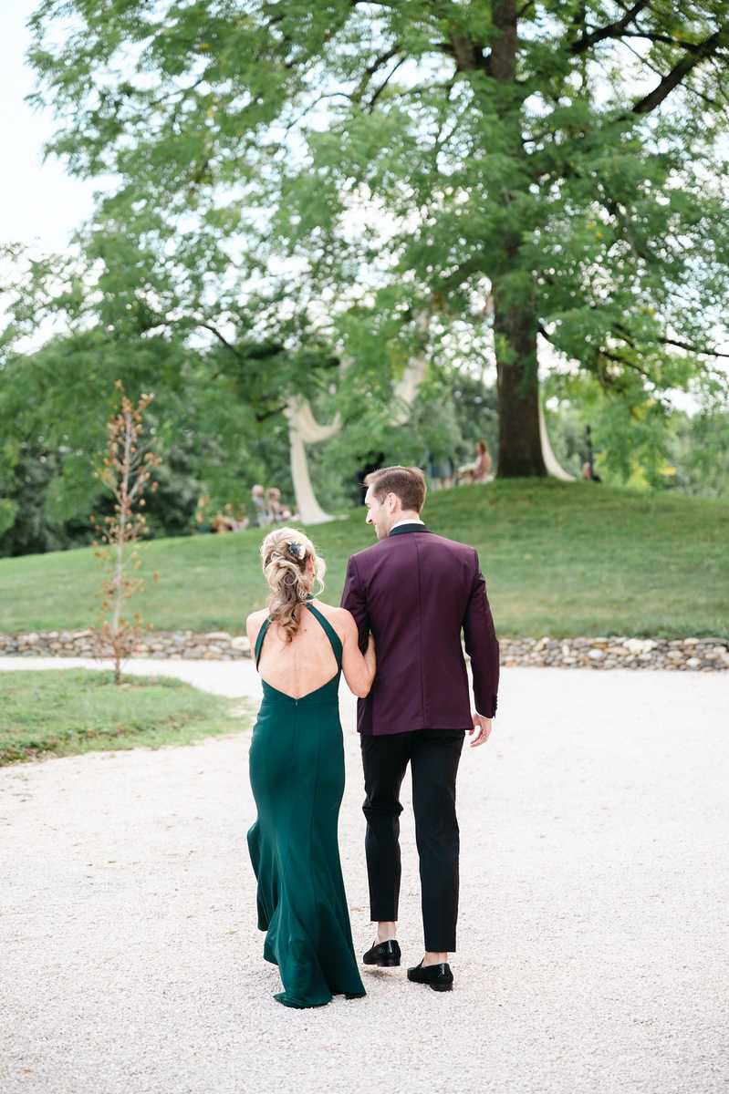 A couple walks arm in arm along a gravel path in an outdoor garden or chateau grounds setting. The woman wears a floor-length forest green halter dress with an open back, her blonde hair styled in a loose updo with a small hair accessory; the man wears a deep burgundy dinner jacket with black trousers and black loafers. The shot is taken from behind, showing both figures walking away from the camera toward a large tree-lined lawn with a low stone wall visible in the background. Other guests can be faintly seen in the distance. The composition is a full-length portrait shot with soft natural light and a slightly blurred background.