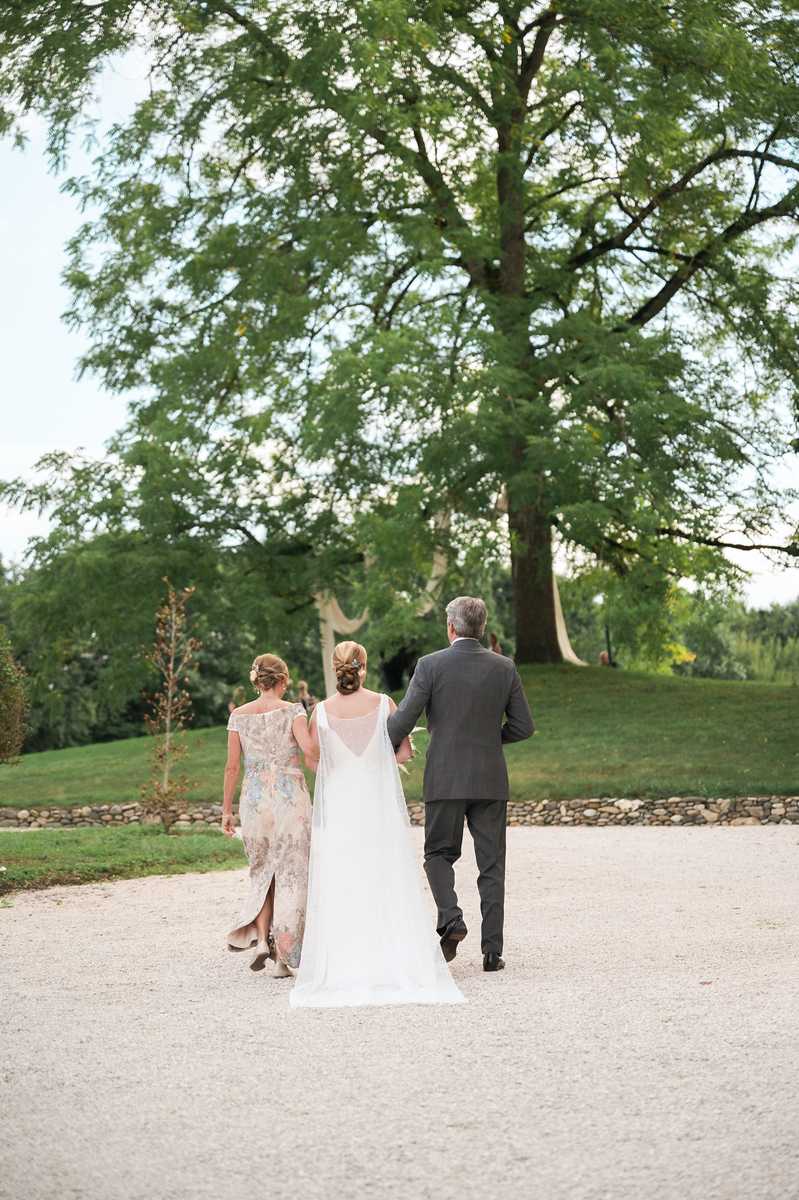 A bride is being walked down a gravel path toward the ceremony by two people, one on each side — a woman in a floral-print off-the-shoulder dress in muted blush and grey tones, and a man in a charcoal grey suit. The bride wears a white sleeveless gown with a sheer low back and a cathedral-length veil, with her hair styled in a low updo with a decorative hair accessory. The shot is taken from behind, showing the trio walking away from the camera toward a landscaped outdoor garden setting with mature trees and a low stone boundary wall. The composition is a wide portrait shot that emphasizes the processional moment and the open outdoor grounds.