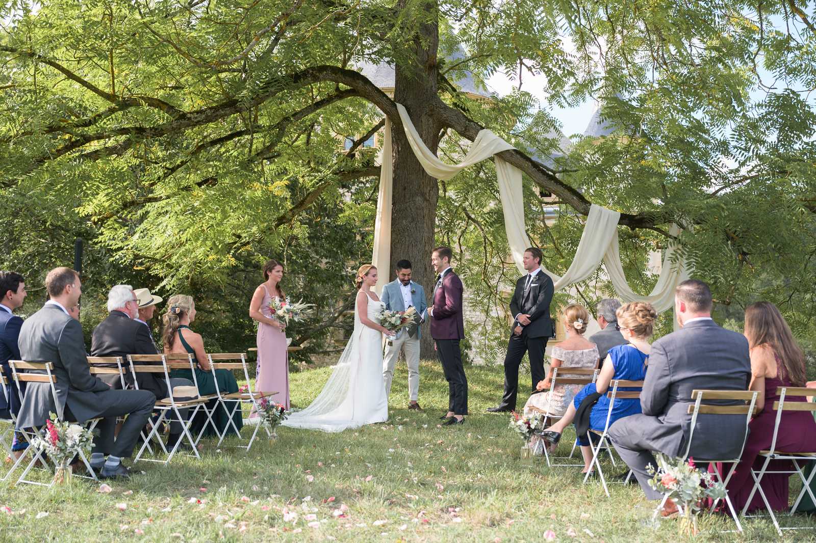 An outdoor wedding ceremony taking place on a lawn in front of what appears to be a chateau, with cream fabric draped between the branches of a large tree serving as the altar backdrop. The bride wears a white sleeveless gown with a long train, holding a loose bouquet of greenery and neutral-toned florals, and stands facing a groom in a light blue suit; a second groom or officiant in a burgundy suit stands beside them, with another man in a dark suit also at the altar. A bridesmaid in a dusty pink sleeveless dress holds a similar greenery bouquet and stands to the left of the couple. Approximately 15–20 seated guests are arranged on white metal folding chairs on either side of a petal-strewn aisle, dressed in a mix of formal attire including cobalt blue, burgundy, and dark green. The ceremony setup has a relaxed, garden-party aesthetic, and the wide shot captures the full scene from a slight distance, showing the aisle, bridal party, officiant, and seated guests in a single frame.