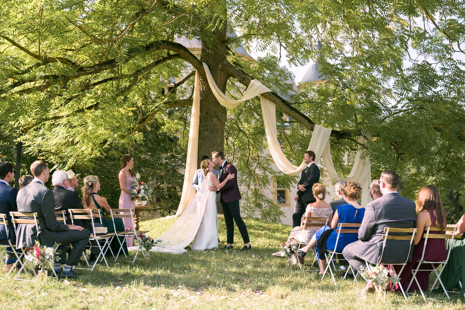 An outdoor wedding ceremony first kiss taking place on a lawn in front of a chateau with a conical-roofed tower visible in the background. The couple stands beneath a large tree decorated with draped ivory fabric serving as the ceremony backdrop, with the bride in a white gown with a long cathedral-length train and the groom in a burgundy suit jacket with black trousers. Two groomsmen in black tuxedos stand to one side and a bridesmaid in a dusty pink dress stands to the other, with small floral arrangements in pink, red, and white with greenery placed at the base of the tree. Approximately 20 guests are seated in white and wooden folding chairs arranged on either side of a grass aisle, dressed in a mix of formal attire including cobalt blue, burgundy, and dark grey. The shot is a wide scene-setting frame capturing the full ceremony layout, altar, and surrounding chateau grounds in bright natural daylight.