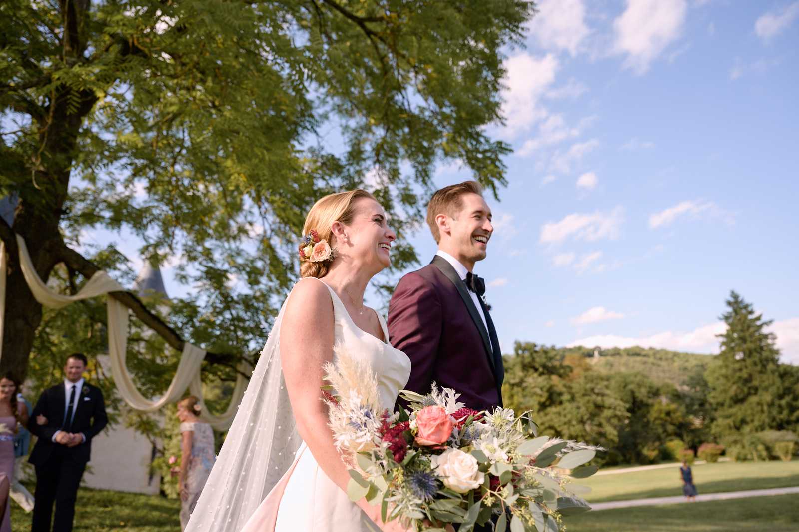 The bride and groom are standing together outdoors during or just after a garden ceremony, both laughing and looking to the side. The bride wears a sleeveless ivory gown with a polka-dot tulle veil and has small peach and pink flowers tucked into her updo; she holds a large loose bouquet of coral roses, deep burgundy blooms, blue thistle, pampas grass, and eucalyptus. The groom wears a deep burgundy tuxedo jacket with black lapels and a black bow tie. In the background, several guests in formal attire are visible near a draped fabric arch, and a chateau turret is partially visible among the trees. The shot is a mid-length portrait taken at a low angle, with the couple positioned left of center against an open lawn and rolling hills.