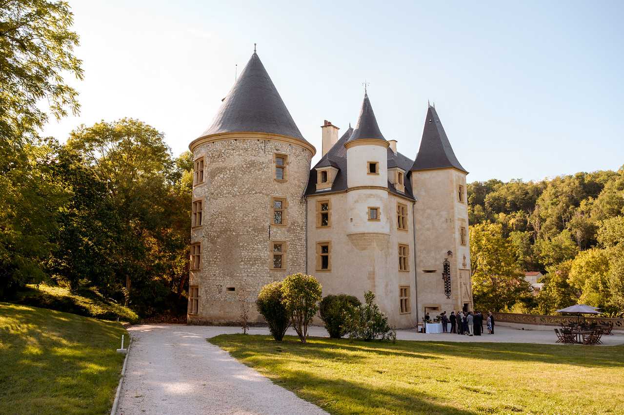 A wide exterior shot of a French château featuring three conical-roofed towers in grey slate, with cream-rendered and exposed stone façades and warm golden-stone window surrounds. A cocktail hour or drinks reception is taking place on the terrace at the base of the château, where approximately 10–12 guests dressed in formal attire are gathered around what appears to be a white-draped drinks or canapé table. To the right, outdoor seating with a grey market umbrella is visible on a paved terrace area. A gravel driveway leads toward the château through a landscaped lawn. Potential venue feature image.