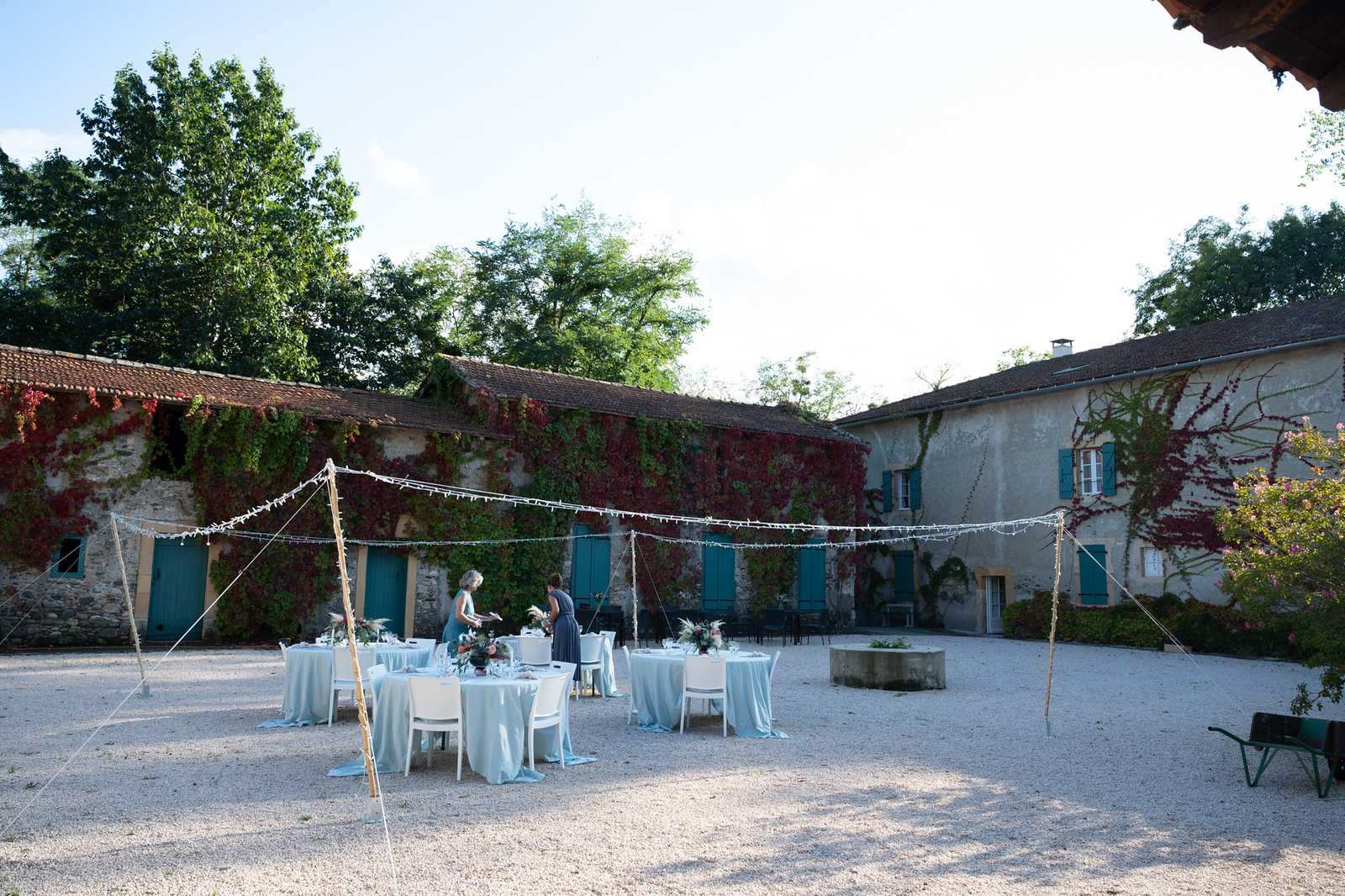 An outdoor wedding reception setup in the gravel courtyard of a French farmhouse or domaine, with stone buildings featuring teal-painted shutters and doors covered in red and green climbing vines forming a U-shape around the space. Several round tables dressed in pale blue-grey linen tablecloths are arranged beneath a canopy of fairy lights strung between bamboo poles, with white chairs and floral centerpieces featuring coral and pink blooms with greenery. Two people, appearing to be event staff or planners dressed in teal and dark grey, are actively arranging the tables in preparation for the reception. The wide shot captures the full courtyard scene including a stone well or fountain feature to the right and a wheelbarrow visible in the foreground corner. Potential venue feature image.