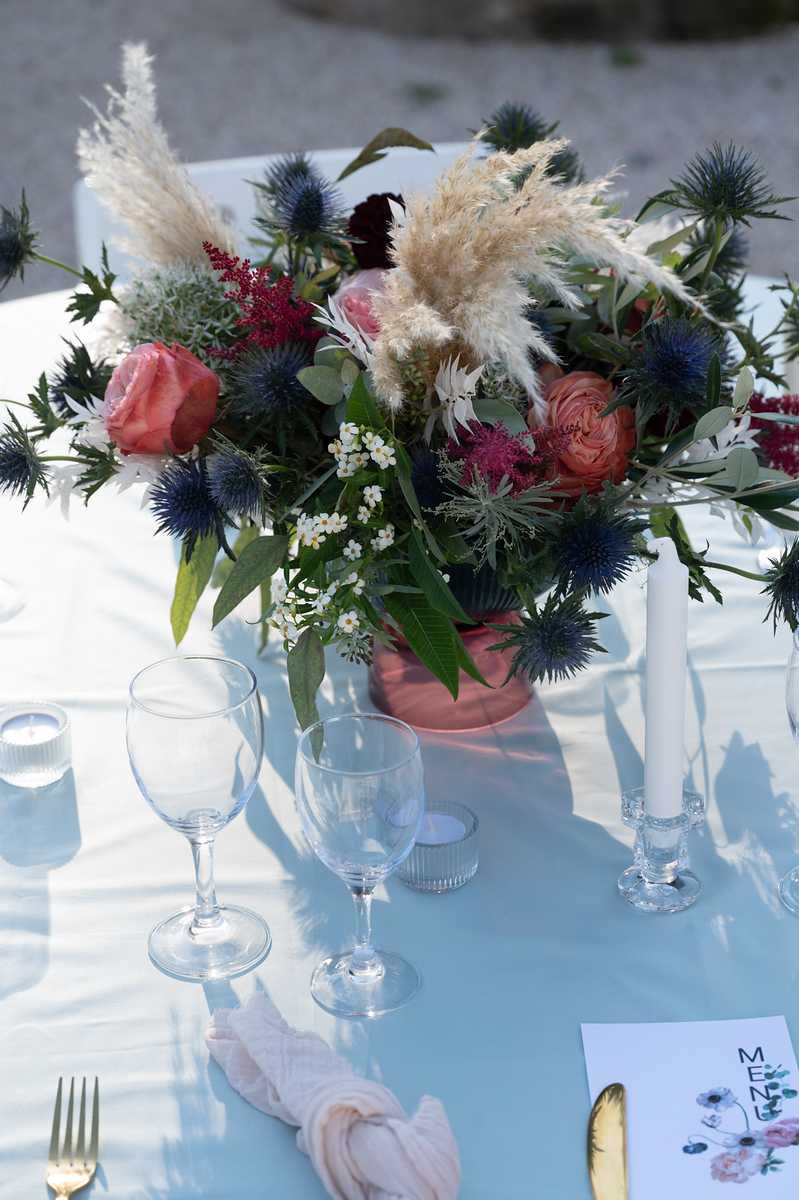 Close-up detail shot of an outdoor wedding reception table set with a pale blue linen tablecloth. The centerpiece is a loose, wildflower-style arrangement in a terracotta-pink vessel featuring coral garden roses, deep burgundy blooms, blue eryngium thistles, pampas grass, astilbe, small white waxflower, and eucalyptus foliage. The table is set with clear wine glasses, small ribbed votive candle holders, white taper candles in glass candlestick holders, gold cutlery, blush pink linen napkins, and a printed illustrated menu card. The overall decor palette combines pale blue, coral, burgundy, and navy in a boho-meets-modern style.