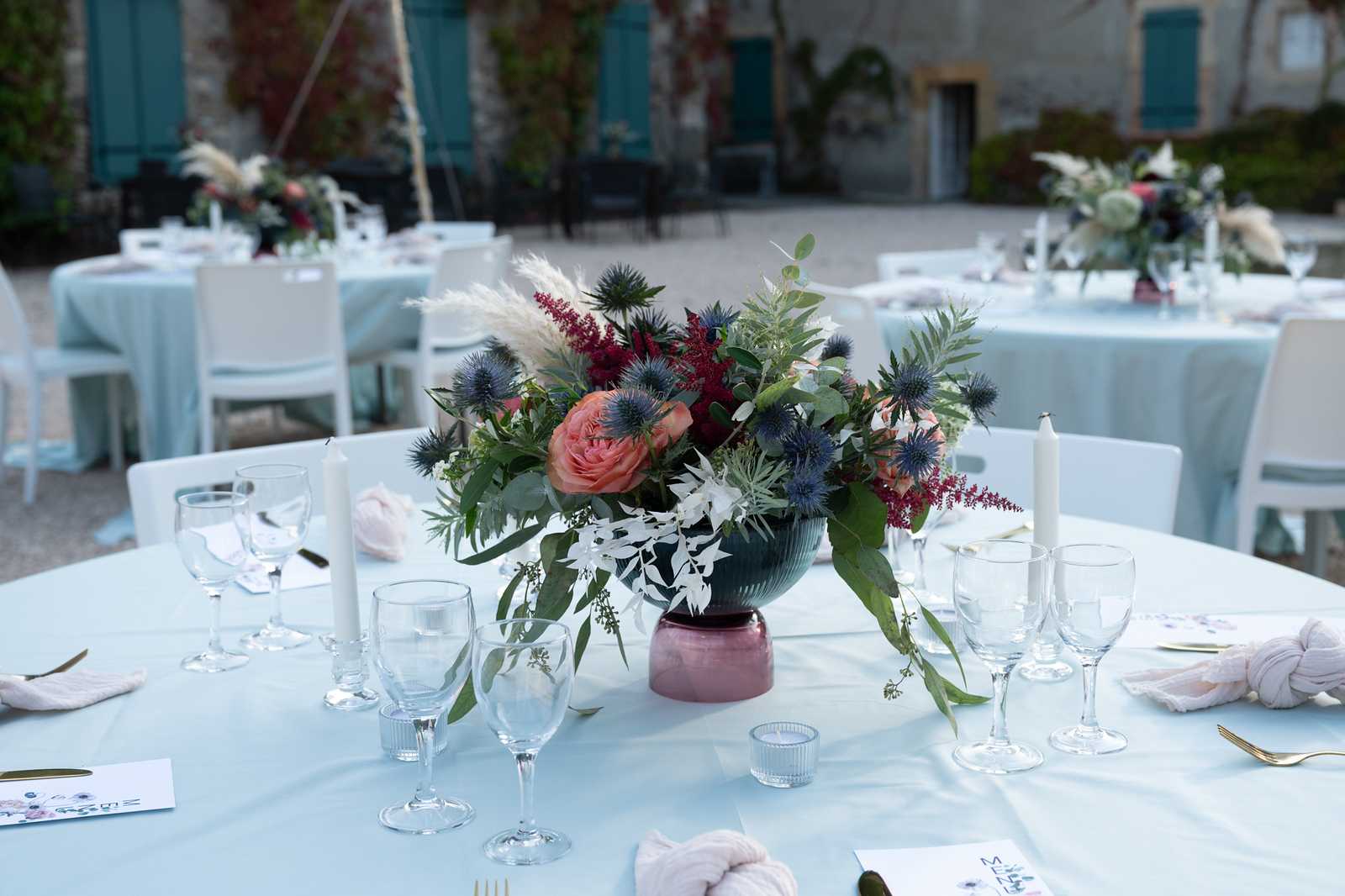 Close-up detail shot of an outdoor wedding reception table setup in a courtyard, with a stone building featuring teal shutters visible in the background. The round tables are dressed in pale powder-blue linen tablecloths and set with clear glassware, white taper candles, small ribbed votive candle holders, blush pink knotted napkins, gold flatware, and printed place cards. The centerpiece is a low, dark teal ribbed bowl with a mauve-pink base, filled with an arrangement of peach garden roses, blue eryngium thistles, burgundy astilbe, white orchids, pampas grass plumes, and trailing eucalyptus foliage. Several additional similarly styled tables are visible in the background, each with matching centerpieces, extending across what appears to be a gravel courtyard. The overall decor palette combines dusty blue, blush, burgundy, and deep teal in a relaxed yet structured style.