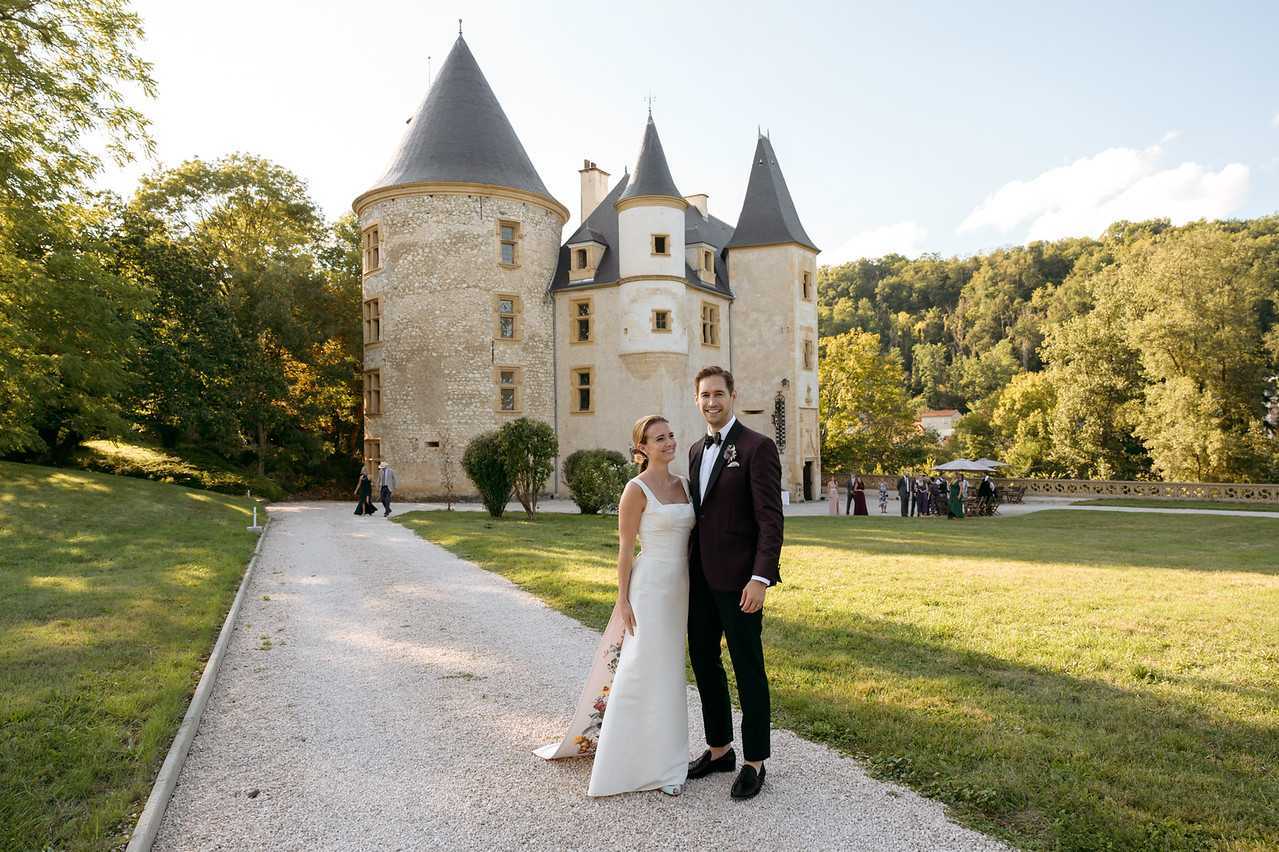 A couple poses for a portrait on a gravel driveway in front of a French château featuring multiple conical-roofed towers and stone façade architecture. The bride wears a sleeveless, column-style white gown with a floral detail visible near the hem, and the groom wears a deep burgundy tuxedo jacket with black trousers, bow tie, and a white boutonnière. The shot is taken during golden hour, with warm late-afternoon light falling across the couple and the château. In the background, a group of approximately 15-20 guests can be seen gathered near a decorative railing on the right side of the grounds, suggesting a cocktail hour in progress. This is a wide portrait shot with the château serving as the primary backdrop. Potential venue feature image.