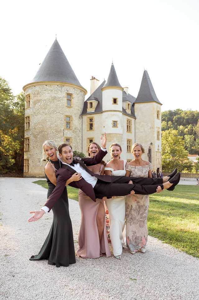 A fun group portrait taken outdoors on a gravel driveway in front of a French chateau featuring multiple conical-roofed stone towers. Four women are carrying a groom horizontally — he is wearing a dark burgundy tuxedo with a bow tie and has his arms spread wide in a playful pose. The group includes the bride in an off-shoulder white gown, a bridesmaid in a dusty rose satin dress, a woman in a dark olive/charcoal floor-length dress, and a woman in a floral off-shoulder gown in muted blush and grey tones. All five are laughing and appear animated. The shot is a medium full-body portrait taken at ground level in natural daylight. Potential venue feature image.