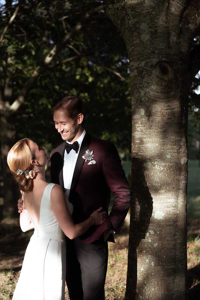 A couple portrait taken outdoors in a wooded setting, with dappled sunlight filtering through the trees. The groom wears a deep burgundy dinner jacket with black lapels, a black bow tie, and a mixed floral boutonniere with small colorful blooms; the bride wears a sleeveless, low-back ivory gown and has her blonde hair styled in an updo adorned with small floral hair accessories. The couple faces each other in a close embrace beside a tree trunk, with the groom smiling down at the bride. The shot is a mid-length portrait with a blurred background of trees and foliage.