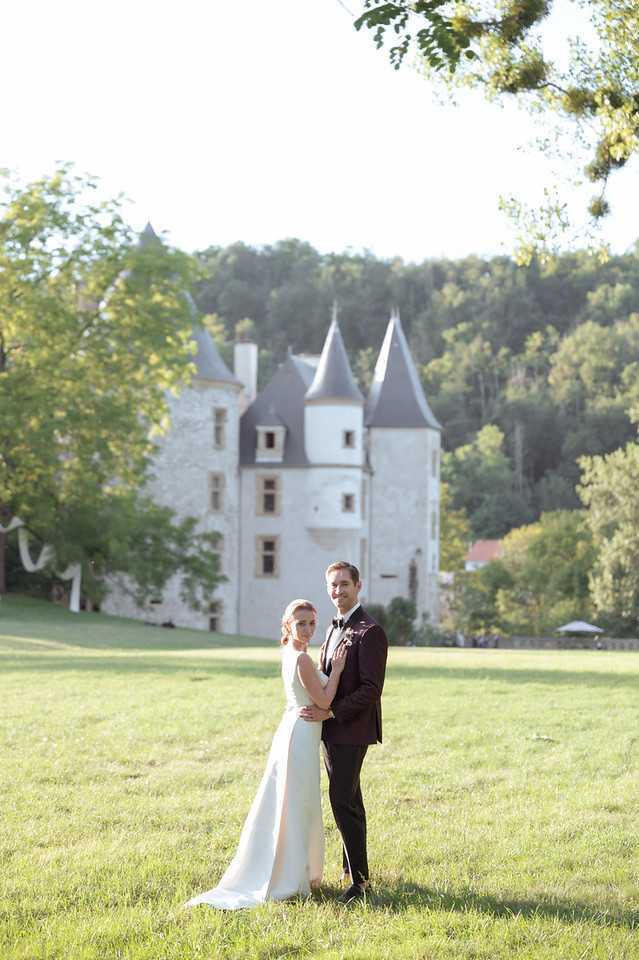 A couple portrait taken outdoors on the grounds of a French chateau, with the white stone castle featuring pointed grey turrets visible in the soft-focus background. The bride wears a sleek, form-fitting ivory gown with a small train, while the groom is dressed in a deep burgundy dinner jacket with black trousers and a black bow tie, with a small boutonniere on his lapel. The two stand closely together on an open lawn, facing the camera in a classic posed portrait composition shot in warm golden-hour light. Potential venue feature image.