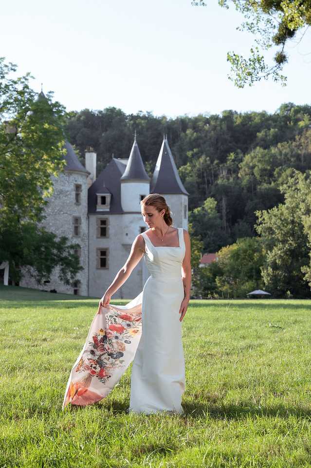 A bride stands alone on the grounds of a French chateau for an outdoor bridal portrait, holding out a detachable train or overskirt with a painted floral print featuring red, orange, coral, and dark navy botanicals on a blush background. She wears a sleek, fitted ivory gown with wide square straps and a minimalist silhouette, with her hair pulled back in a low updo. The chateau in the background is a classic French castle with grey slate conical turret roofs and pale stone walls. The shot is a full-length portrait taken at a medium distance with the chateau centered behind her as a backdrop. Potential venue feature image.