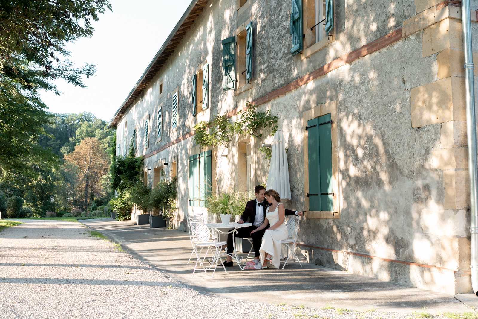 A bride and groom sit together at a small white wrought-iron bistro table and chairs along the exterior facade of a large French country estate or domaine, sharing a quiet moment during what appears to be a couples portrait session. The groom wears a black tuxedo with a bow tie, and the bride wears a sleek ivory sleeveless gown with pink sneakers visible at her feet. The building features aged rendered stone walls, teal-green shutters, and brick detailing, with climbing plants framing the windows. The wide shot captures the full length of the building's facade, a gravel driveway, and dappled tree shade falling across the couple and the wall behind them. Potential venue feature image.