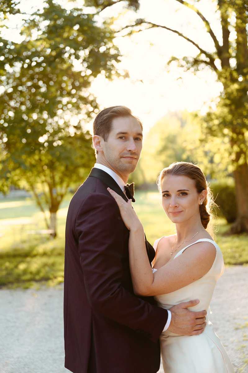 Outdoor couple portrait taken during golden hour, with warm backlit sunlight creating a bright, glowing background. The groom wears a deep burgundy tuxedo jacket with a black bow tie, and the bride wears an ivory sleeveless gown with thin straps, her dark hair pulled back in a low braid, and a delicate pendant necklace. She stands behind him with her arms wrapped around his torso, both turning their faces toward the camera with calm expressions. The setting appears to be a gravel path lined with mature trees on a chateau or estate grounds. The composition is a close-up portrait with a shallow depth of field, softly blurring the background.