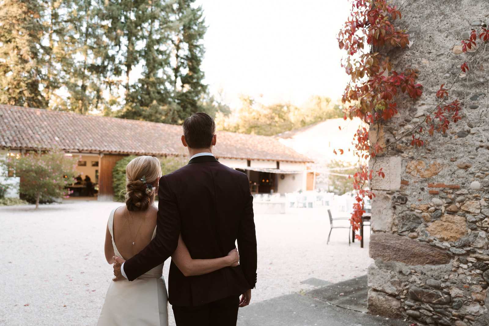 A bride and groom are photographed from behind as they walk arm-in-arm through a gravel courtyard of a French country estate, viewed at golden hour. The bride wears a white gown with a low open back, her blonde hair styled in a low chignon with a small floral hair accessory, while the groom wears a dark navy suit. The venue features low stone buildings with terracotta tile roofs, a rustic wooden barn structure with warm interior lighting visible in the background, and a textured stone wall to the right draped in deep red climbing vines. The composition is a wide portrait shot taken from behind, with the couple framed between the stone architecture and the softly lit courtyard beyond. Potential venue feature image.