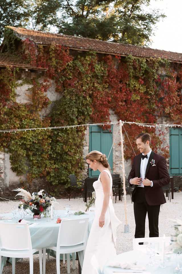 The bride and groom stand together at an outdoor reception in a courtyard, appearing to look over the table setup. The bride wears a sleeveless ivory A-line gown with an updo hairstyle, while the groom wears a dark navy or black tuxedo jacket with a black bow tie and a floral boutonniere. The reception tables are dressed with pale blue-gray linen and feature low centerpieces of mixed autumnal florals including pampas grass, burgundy and orange blooms, and thistle accents. String lights are strung overhead between posts, and the backdrop is a stone outbuilding covered in autumn ivy in deep reds and greens with teal-painted shutters. The shot is a medium portrait taken from a slight distance, capturing both the couple and the table decor in context.