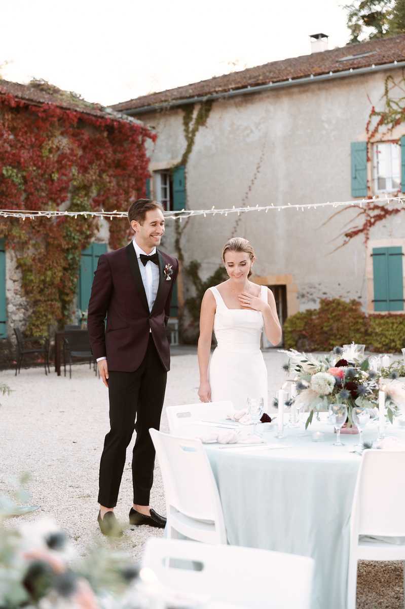 A couple stands beside a decorated reception table in the courtyard of a French country property, appearing to see the table setup for the first time. The groom wears a deep burgundy tuxedo jacket with black trousers, a bow tie, and a small floral boutonnière, while the bride wears a fitted white square-neck sleeveless dress with her hair pulled back. The round table is dressed with a pale blue-grey linen and features a low centerpiece of white hydrangeas, deep burgundy dahlias, dusty blue thistles, and trailing greenery, with crystal glassware and white chair covers visible. Fairy lights are strung overhead across the courtyard, and the venue building in the background has teal-painted shutters. The shot is a mid-length portrait taken outdoors in natural light.