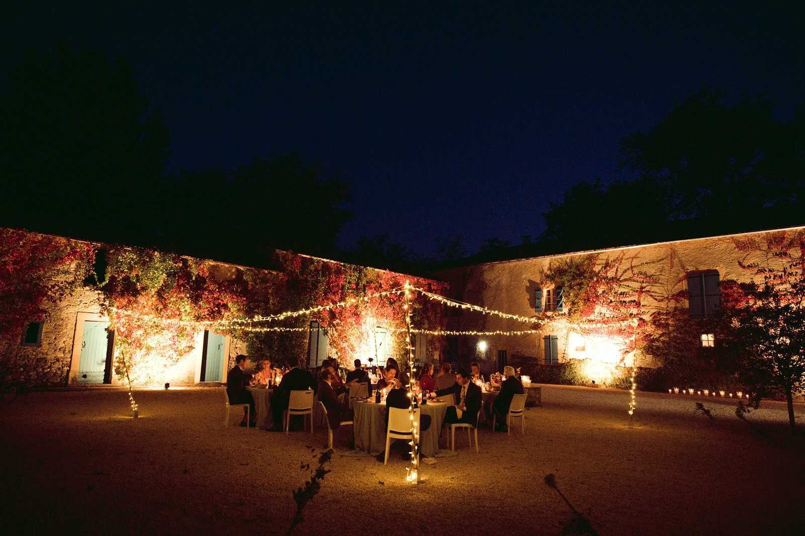 An outdoor evening wedding reception taking place in a courtyard of a French stone-walled property, likely a domaine or mas. Approximately 20–25 guests are seated at several round tables covered with grey linen, arranged on a gravel courtyard enclosed by two wings of the building. Fairy lights are strung across the courtyard on poles, and small candles line the perimeter walls, creating warm amber lighting against the night sky. The stone walls are heavily covered in climbing vines displaying deep red and green autumn foliage, which is dramatically uplit, adding rich color to the scene. Light blue-grey shuttered doors and windows are visible along the building facades. The overall styling is rustic and autumnal, with warm candlelit tones contrasting the deep blue night sky. Wide establishing shot capturing the full courtyard setting. Potential venue feature image.