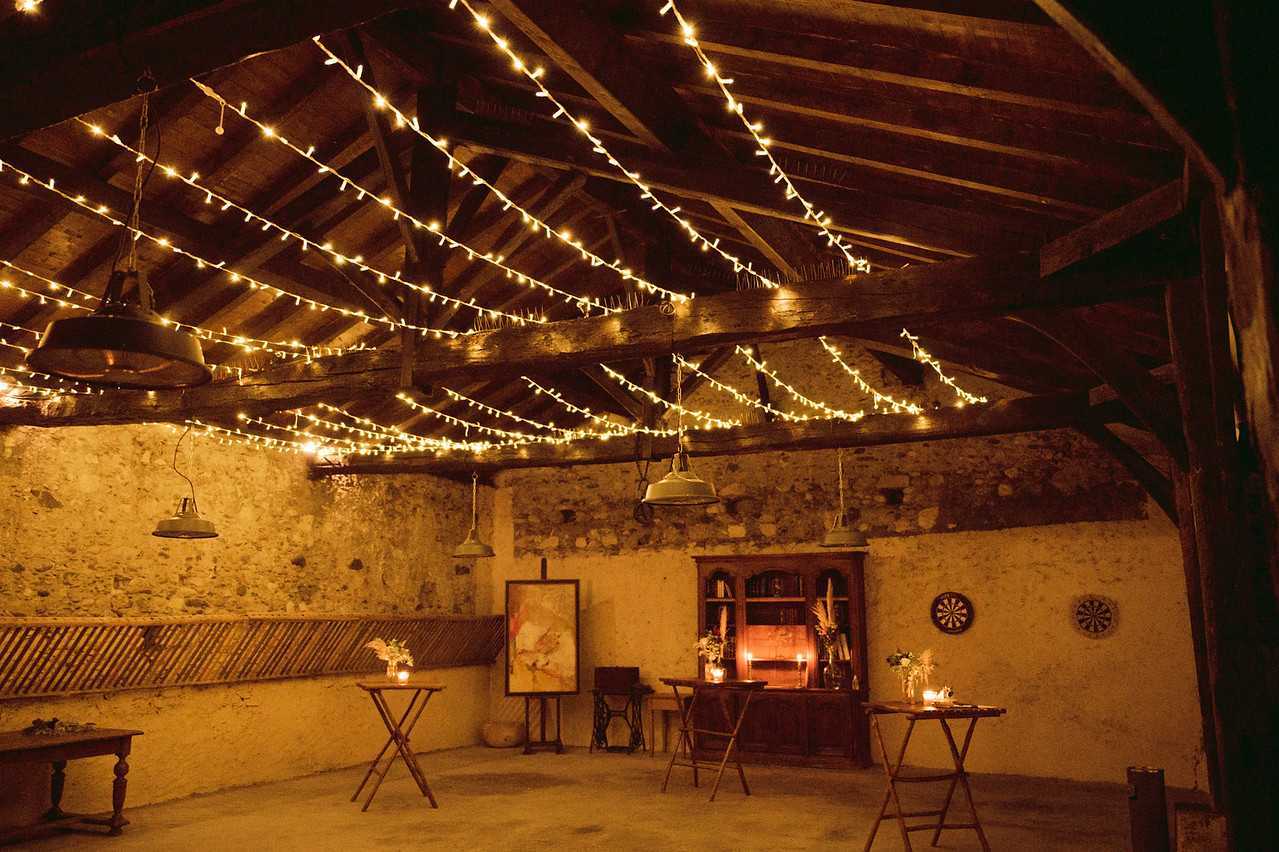 An indoor reception or cocktail space set up inside a rustic barn or stone outbuilding, photographed as a wide interior shot with no people present. The exposed wooden beam ceiling is densely draped with warm white fairy lights strung in diagonal crisscross patterns, while several vintage-style industrial enamel pendant lights hang from the beams. The walls are bare stone and plaster, adding to the rustic aesthetic. The space is furnished with several folding trestle tables holding small floral arrangements and candles, a wooden bookcase unit lit from within by candles, a framed artwork on an easel, and two dartboards mounted on the far wall. The overall decor palette is warm amber and gold, driven entirely by candlelight and fairy light illumination, consistent with a rustic or barn-style wedding theme. Potential venue feature image.