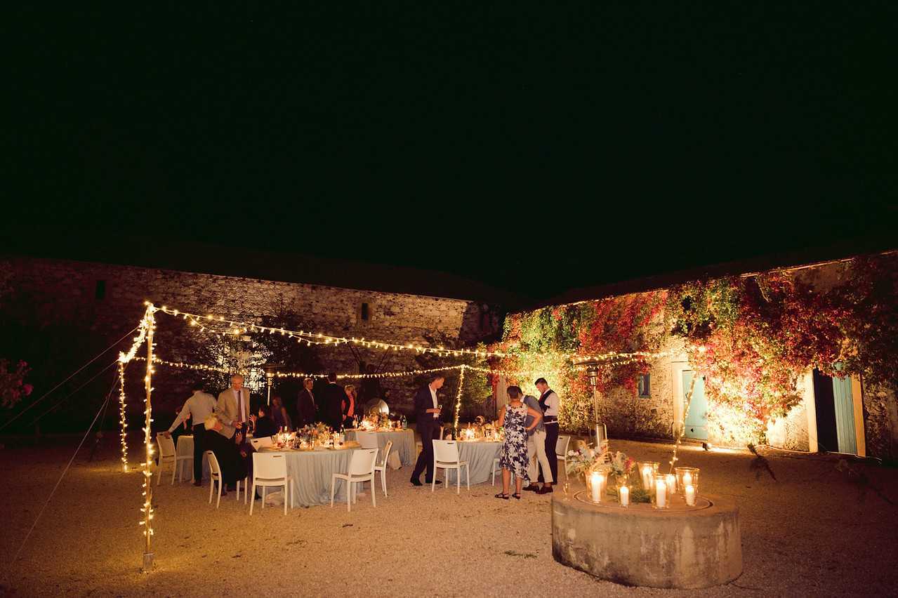 An outdoor evening wedding reception taking place in a courtyard surrounded by old stone buildings, with creeper vines covering one wall lit in warm amber and red tones. Approximately 15 guests are gathered around round tables dressed in pale blue-grey linen tablecloths, set with white chairs, candles, and floral centerpieces. The space is lit by strands of festoon fairy lights strung overhead between wooden poles, creating a warm, intimate atmosphere. In the foreground, a low circular stone feature holds a cluster of pillar candles in varying heights. The overall styling is rustic with a relaxed, convivial mood, captured in a wide shot that shows the full courtyard setting at night.