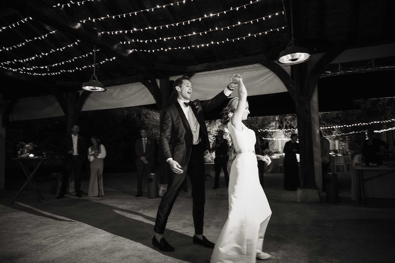 A black-and-white image of the couple's first dance at an outdoor evening reception under a wooden pergola structure. The groom, wearing a dark tuxedo with a bow tie, is spinning the bride, who wears a sleek sleeveless white gown; both are laughing and visibly joyful. Multiple strands of bistro/globe string lights are draped across the pergola ceiling, with pendant industrial-style lights also hanging from the structure, and additional string lights visible along the surrounding walls in the background. Approximately six to eight guests can be seen watching in the background, with candlelit reception tables visible to the sides, suggesting a classic rustic-chic outdoor reception setting. The image is captured in a wide portrait orientation with high contrast black-and-white tones, giving the scene a dramatic, deep-shadow quality.