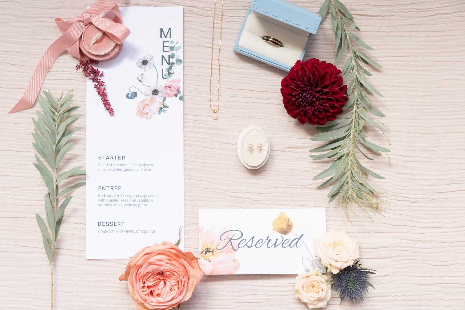 A flat lay detail shot of wedding stationery and accessories arranged on a light wood surface. The centerpiece is a printed menu card featuring a watercolor floral design with anemones and peonies in blush and white tones, listing a three-course meal of starter, entrée, and dessert, alongside a watercolor 'Reserved' place card. Accessories include a diamond engagement ring in a dusty rose velvet ring box with a blush satin ribbon, a gold band in a powder blue ring box, pearl stud earrings in a cream oval jewelry holder, a delicate gold chain necklace with a small pearl pendant, and a cluster of eucalyptus branches. Florals include a deep crimson dahlia, a peach garden rose, cream spray roses, and a steel blue thistle, with small burgundy dried botanicals adding texture to the arrangement.