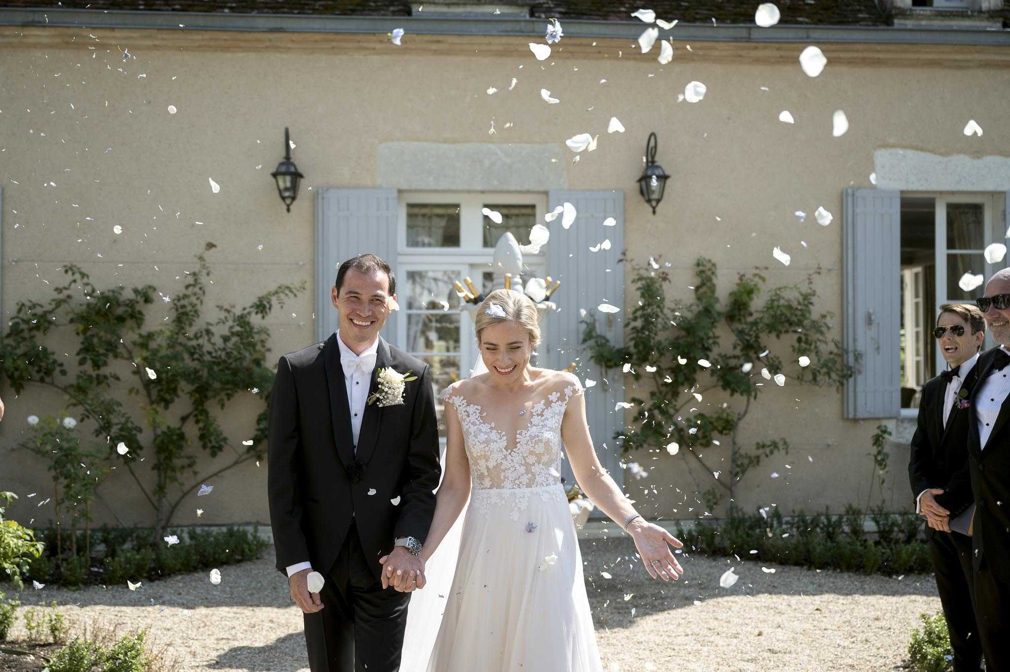 The bride and groom walk hand-in-hand through a shower of white rose petals thrown by guests during their post-ceremony exit, outdoors on a gravel path in front of a pale yellow French country manor or château with grey-blue shutters. The bride wears a cap-sleeve gown with an illusion lace bodice over a nude underlay and a flowing ivory skirt, with her hair pinned up and adorned with a small floral piece; the groom wears a black tuxedo with a white bow tie and a small white floral boutonnière. Both are laughing, and at least two male guests in black tuxedos are visible on the right edge of the frame, also reacting with laughter. The shot is a medium wide portrait taken in bright natural daylight, with petals frozen mid-air across the full frame.