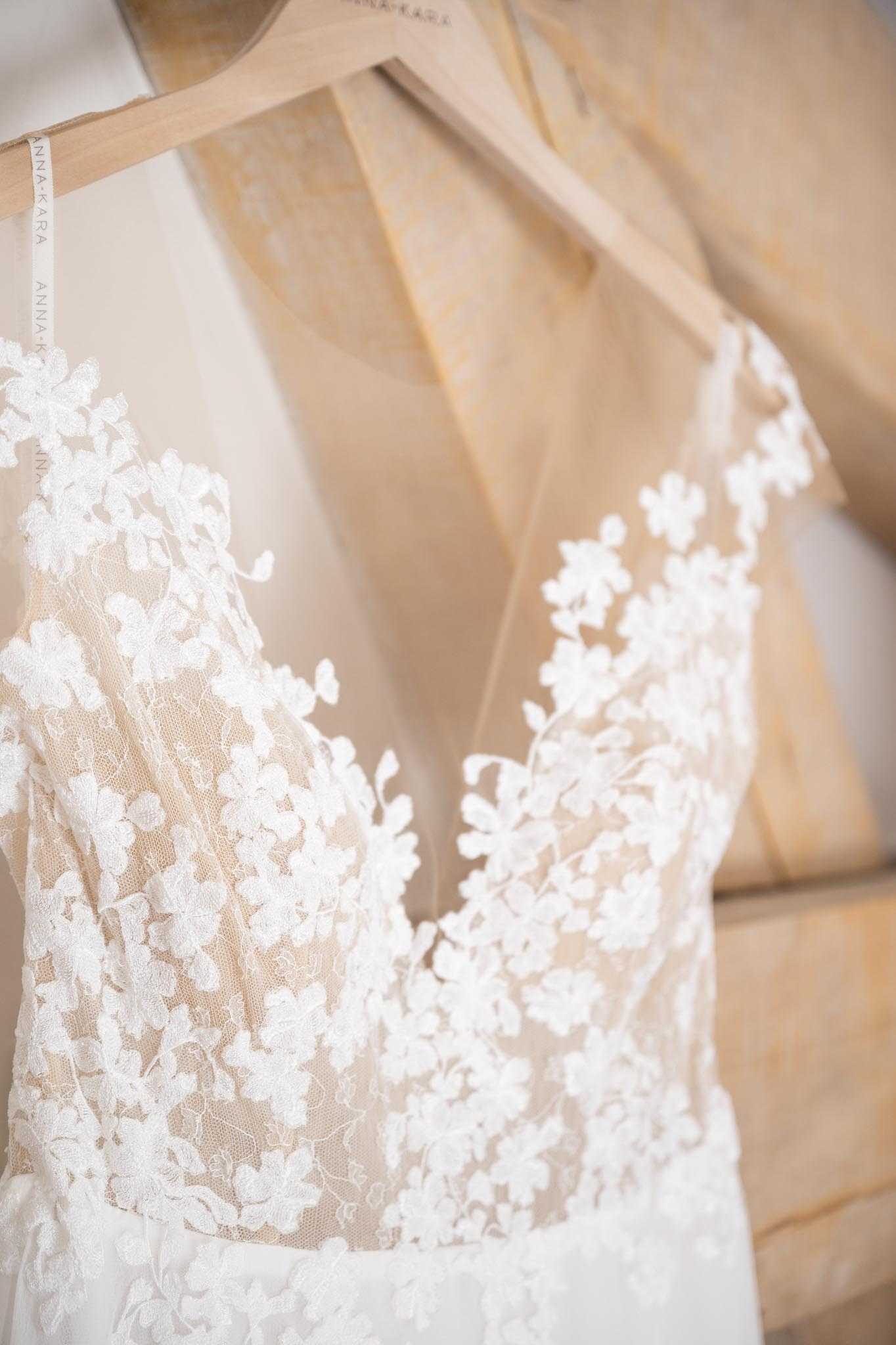 Close-up detail shot of a wedding dress hanging on a natural wood hanger, taken during the getting-ready phase. The gown is by Anna-Kara, as visible on the hanger label, and features a deep V-neckline with ivory three-dimensional floral lace appliqués layered over a nude/champagne illusion tulle bodice. The lace detailing includes small clustered flowers and trailing leaf motifs, creating a romantic, delicate texture across the sheer upper bodice. The background is softly blurred, showing warm neutral tones consistent with an indoor preparation setting.