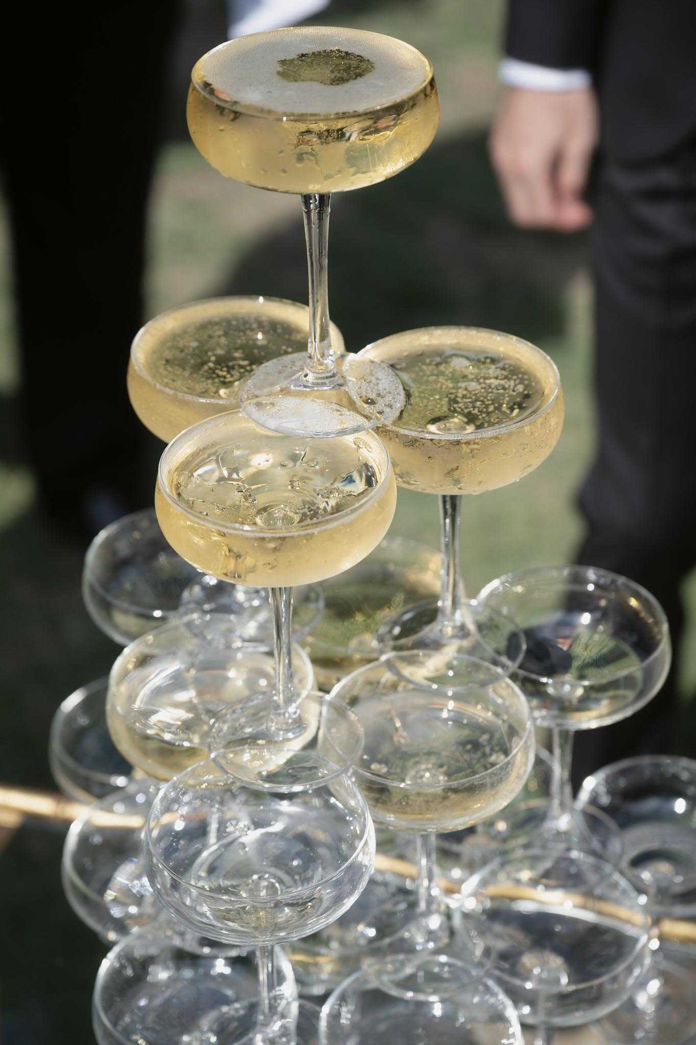 Close-up detail shot of a champagne tower built from coupe glasses stacked in a pyramid formation. The upper tiers are filled with pale golden champagne showing visible bubbles and foam, while the lower tiers remain empty, suggesting the pour is in progress or yet to begin. A person dressed in a dark suit is partially visible in the blurred background, indicating this is likely during a cocktail hour or reception. The coupe-style glassware and champagne tower format reflect a classic French wedding reception tradition.