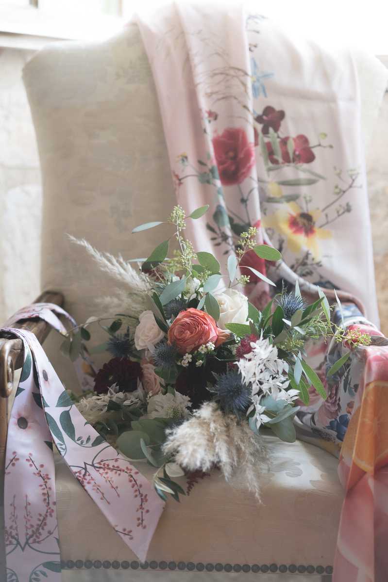 Close-up detail shot of a bridal bouquet resting on an upholstered armchair draped with a floral-printed pink fabric. The bouquet features coral garden roses, ivory roses, blush ranunculus, deep burgundy dahlias, blue thistle, white astrantia, wax flower, eucalyptus, and pampas grass plumes, giving it a loose, garden-style boho aesthetic. The bouquet is tied with a blush pink ribbon printed with a botanical leaf and berry motif. The overall color palette mixes coral, burgundy, dusty blue, ivory, and sage green against the soft pink floral textile backdrop.