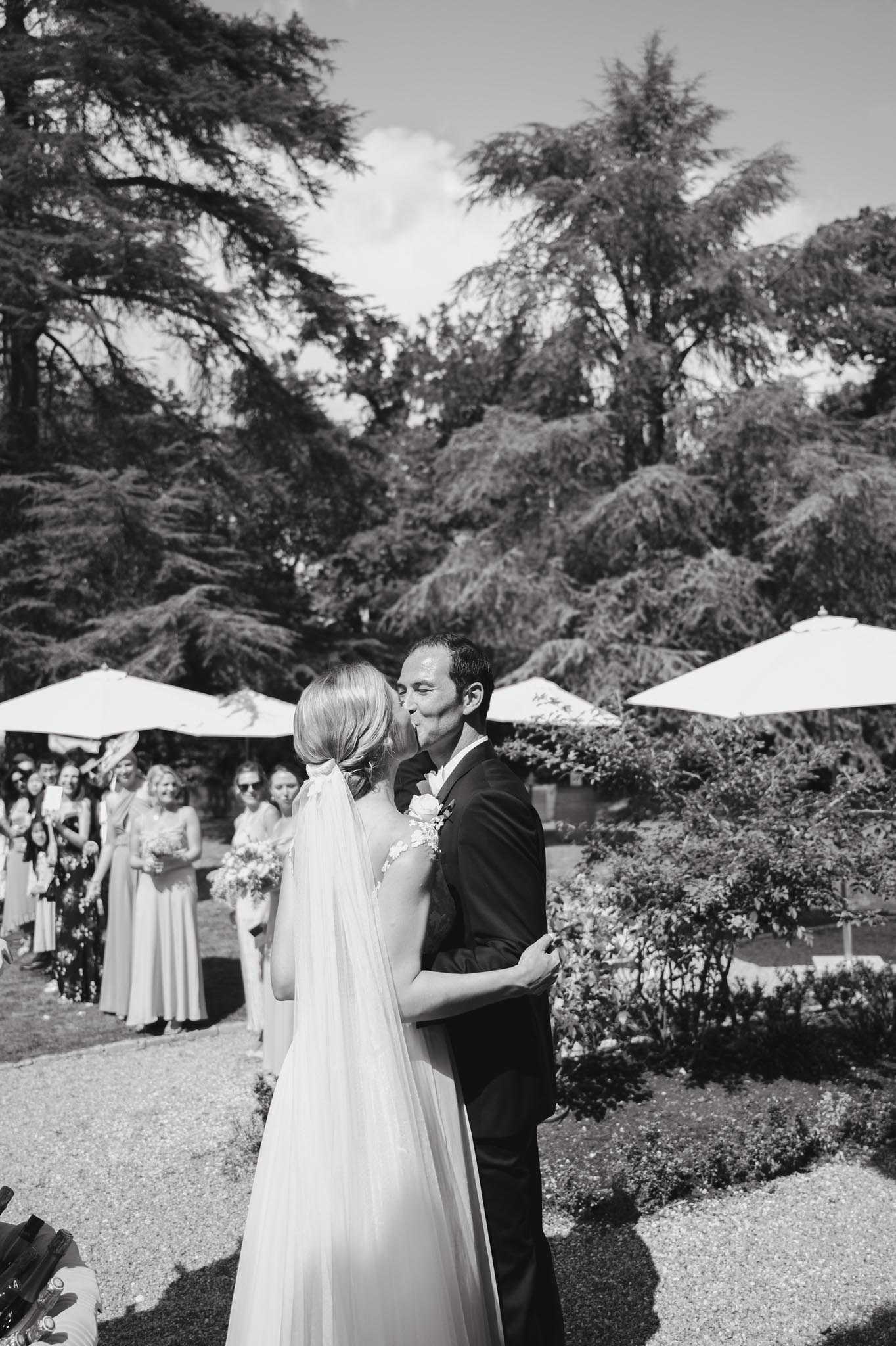 This black-and-white image captures the couple's first kiss during an outdoor ceremony on a gravel area within a formal garden setting. The bride wears a flowing gown with a long veil and has her hair up, while the groom is dressed in a dark suit with a floral boutonnière. In the background, approximately 10–15 guests look on, several of them holding small bouquets, with white market umbrellas visible among them. The shot is a mid-range portrait taken from behind the bride, giving a slightly candid, documentary feel with strong contrast between the bright sky and the dense dark foliage of large cedar trees overhead.
