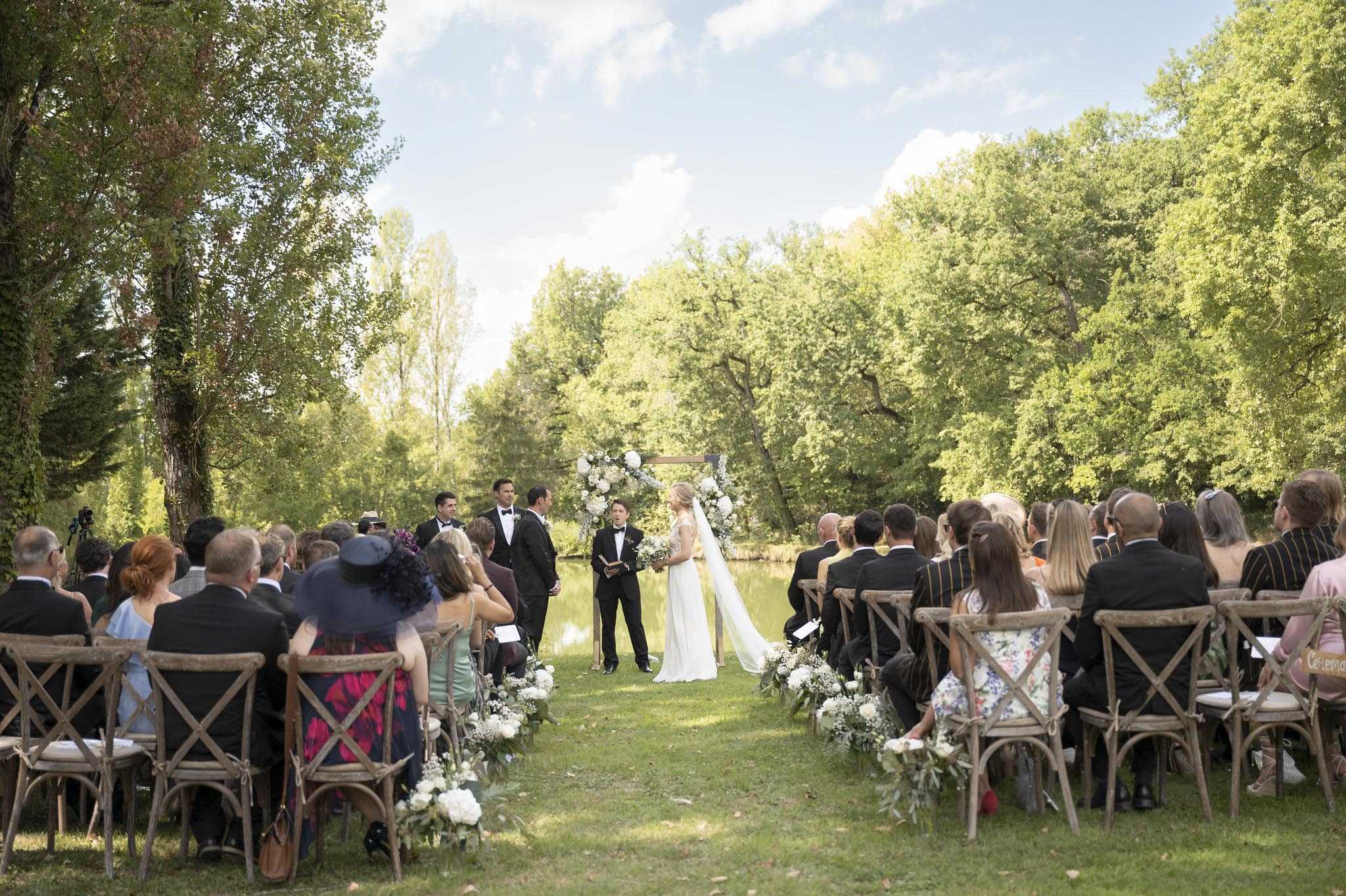 An outdoor wedding ceremony taking place on a lawn beside a calm river or pond, with a dense tree canopy forming the backdrop. The bride, wearing a white gown with a long cathedral veil and carrying a white bouquet, stands facing two grooms in black tuxedos with bow ties, while an officiant in a black suit reads from a book at the altar. The altar is framed by a wooden arch adorned with large white floral clusters, and the grass aisle is lined on both sides with low arrangements of white flowers — including what appear to be hydrangeas, peonies, and baby's breath with green foliage. Approximately 60–80 guests are seated in natural wood cross-back chairs arranged in two sections, dressed in formal and semi-formal attire. The overall decor palette is white and green with a classic, garden-party aesthetic, and the image is taken from a wide, mid-distance shot looking down the aisle toward the altar.