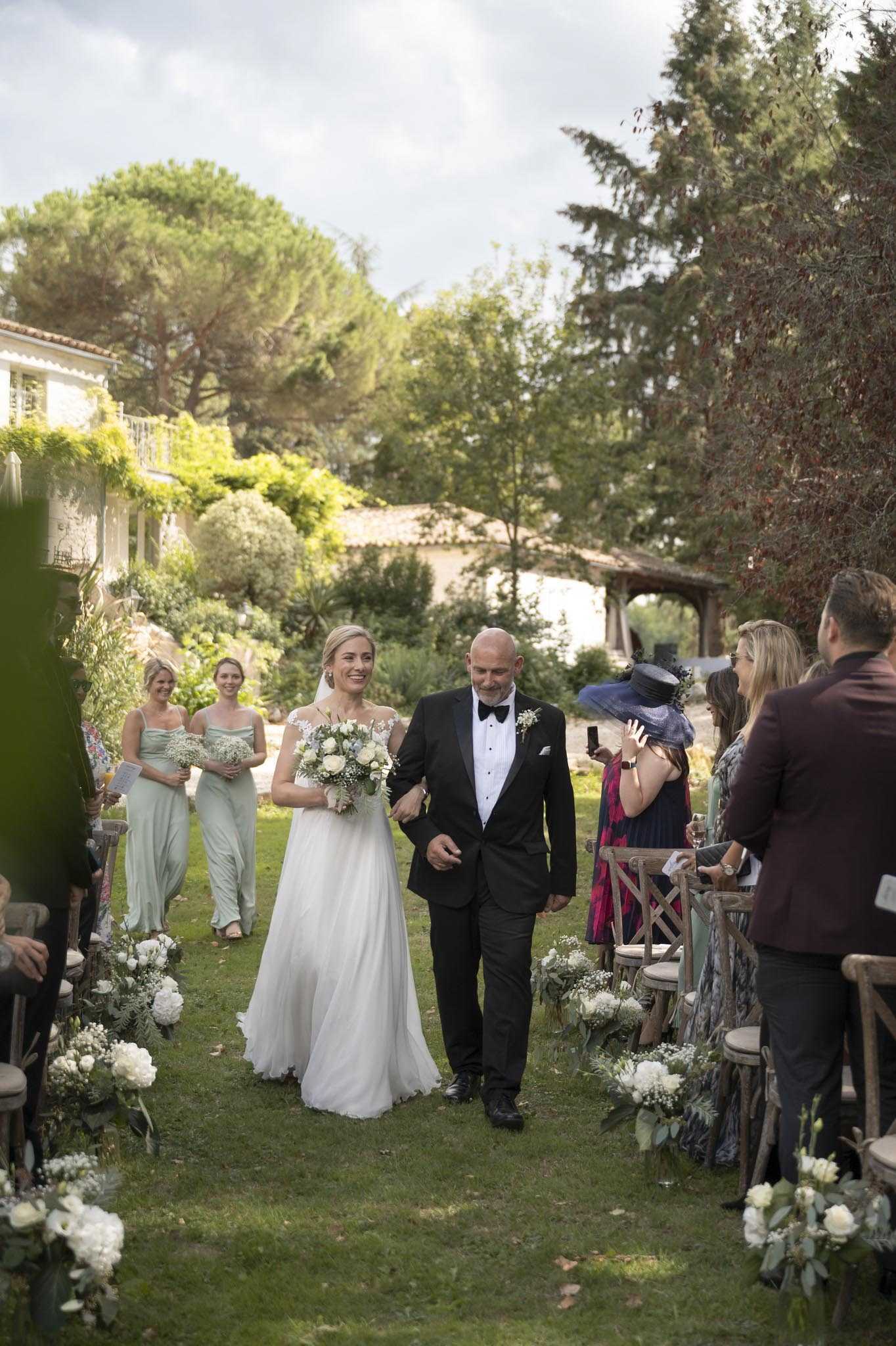 The bride is being walked down the aisle during an outdoor garden ceremony at what appears to be a Provençal stone mas or country house. She wears a flowing white gown with lace cap sleeves and carries a loose bouquet of white roses, white peonies, and baby's breath, while her escort wears a classic black tuxedo with bow tie and a white floral buttonhole. Two bridesmaids follow behind in sage green floor-length dresses, each holding small bouquets of baby's breath. The grass aisle is lined on both sides with clusters of white flowers — including white roses, peonies, and baby's breath — arranged at ground level alongside rustic wooden cross-back chairs. Seated and standing guests are visible on either side, including a woman in a navy wide-brim hat. The overall decor palette is white and sage green with a classic garden-party aesthetic. Wide shot taken from a low angle at the end of the aisle looking toward the couple.