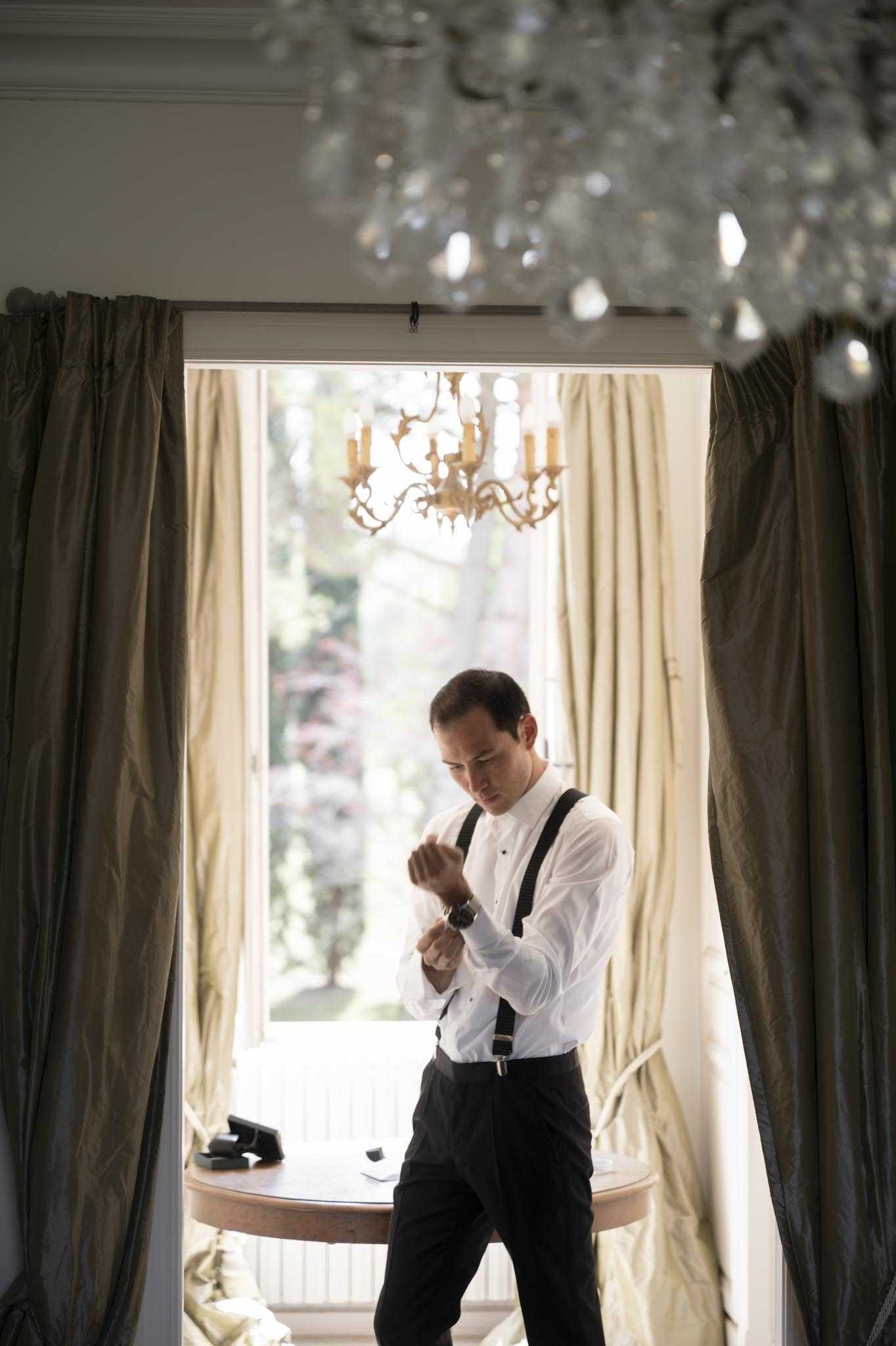 A groom is getting ready indoors, fastening his cufflinks or watch while standing in front of a large window that floods the room with natural light. He is wearing a white tuxedo shirt with black suspenders and black trousers, with a watch visible on his wrist. The room features sage-green silk floor-length curtains, a crystal chandelier in the foreground, and a gold ornate chandelier visible through the window, suggesting a classic French chateau or manor house interior. The composition is a medium full-length portrait with the groom backlit by the window, creating a warm, softly contrasted atmosphere.