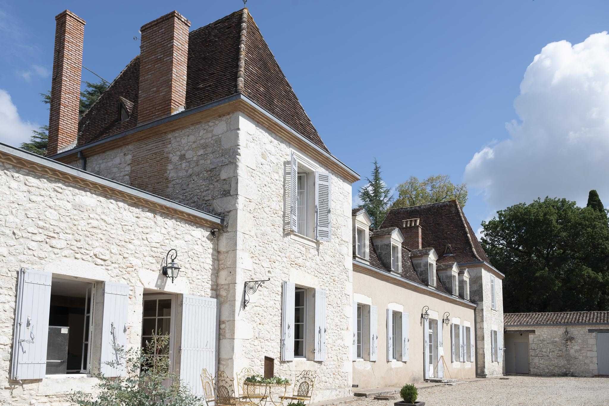 Wide exterior shot of a French country estate featuring cream-toned limestone buildings with pale grey-blue shutters, terracotta tile roofs, and tall brick chimneys. The property includes a main two-storey tower section connected to a lower wing, with a second manor building visible behind it to the right. A gravel courtyard sits in the foreground with wrought iron gold bistro chairs and a small table positioned near the entrance. Wall-mounted iron lanterns flank the doorways, and a pair of clipped box topiary balls sit near the lower building's entrance. No people are present in the image. Potential venue feature image.