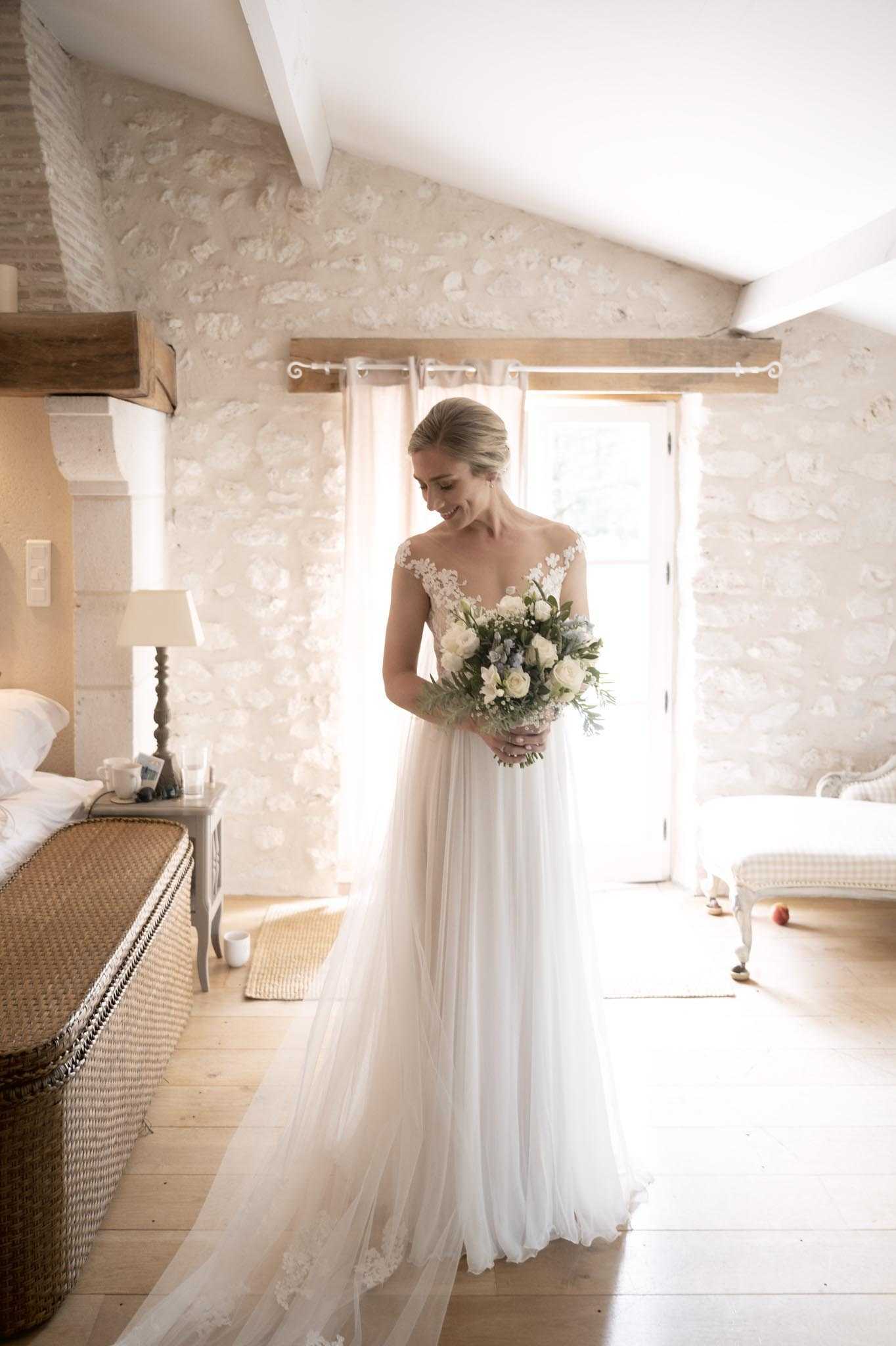 A bride stands alone in a getting-ready portrait inside a rustic French château bedroom with exposed limestone walls, wooden ceiling beams, and wide-plank oak floors. She wears an ivory tulle A-line gown with an illusion lace cap-sleeve bodice and a flowing skirt with a train, her blonde hair pulled back in a low updo. She holds a loosely gathered bouquet of white roses, white freesia, dusty blue accents, and trailing greenery including eucalyptus, and looks downward with a smile. The room features a grey bedside table with a lamp, sheer blush curtains, a wicker bed frame, and a French provincial upholstered stool, giving the space a relaxed classic style; the shot is a full-length portrait framed by natural backlight from an open French door.