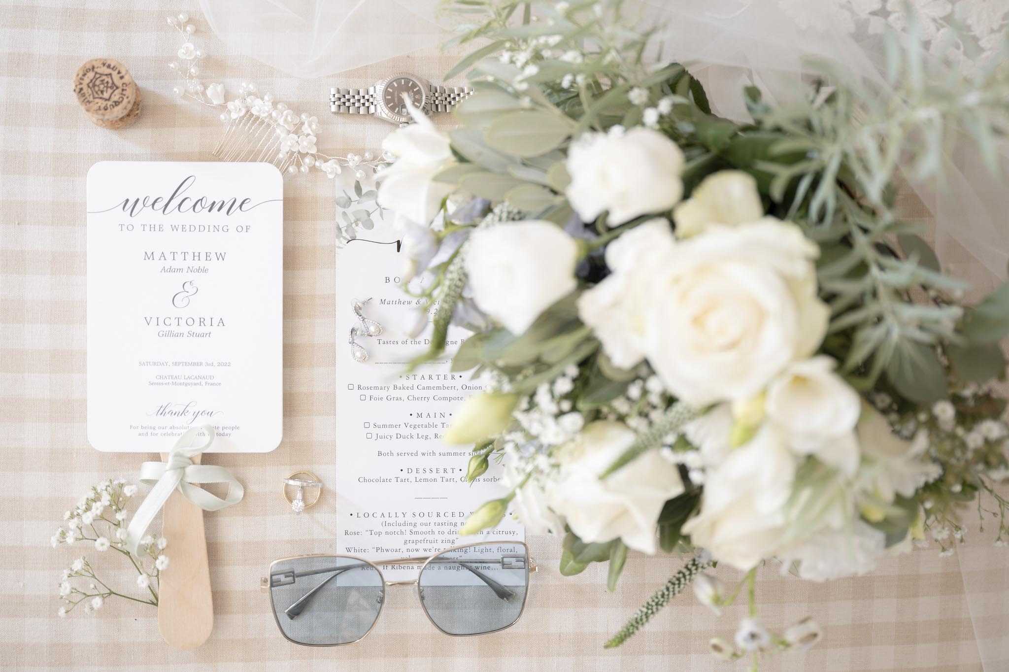 A flat lay detail shot arranging wedding stationery and accessories on a beige gingham-patterned fabric surface. The items include a white wedding program reading 'Welcome to the Wedding of Matthew Adam Noble & Victoria Gillian Stuart, Saturday September 3rd, 2022, Chateau Lacanaud, Sencas-et-Montpézard, France,' a partially visible menu card listing starters of Rosemary Baked Camembert and Foie Gras, a bouquet of ivory garden roses, white lisianthus, baby's breath, and soft green eucalyptus foliage, a silver Rolex watch, drop crystal earrings, a gold ring, a pearl and crystal hair comb, a wine cork, a pale sage ribbon bow, and a pair of silver-framed sunglasses with grey-tinted lenses. The color palette is consistent throughout — white, ivory, sage green, and silver — reflecting a classic, refined styling theme.