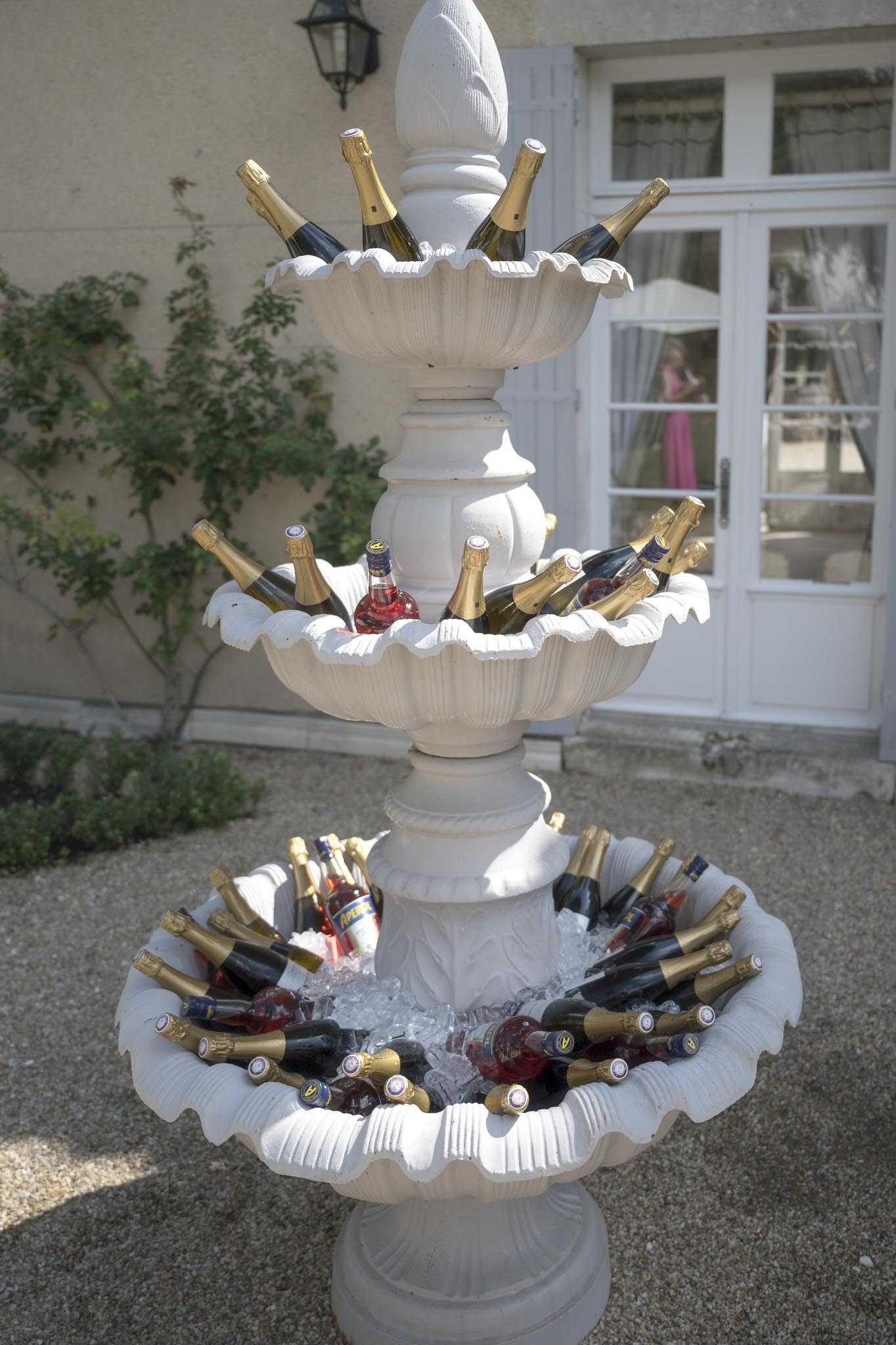 A cocktail hour detail shot featuring a large three-tiered white stone fountain repurposed as a drinks display, filled with bottles of champagne with gold foil tops, rosé wine, and what appear to be Aperol bottles, all chilled on ice. The setup is outdoors on a gravel courtyard in front of a classic French manor-style building with white-shuttered French doors and a wall-mounted lantern. The styling is classic and celebratory, with the fountain serving as a dramatic centerpiece for drink service. In the background, a person in a pink dress is visible through the glass doors of the building.