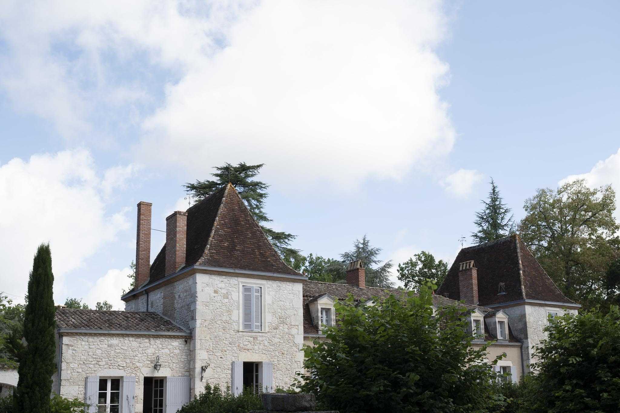A wide exterior shot of a traditional French country manor house, featuring cream-colored stone facades, dark brown tiled steeply pitched rooftops with dormer windows, white-shuttered windows, and brick chimneys. The building spans multiple connected wings with conical tower rooflines typical of French regional architecture. No people or wedding decor are visible in this image. Potential venue feature image.