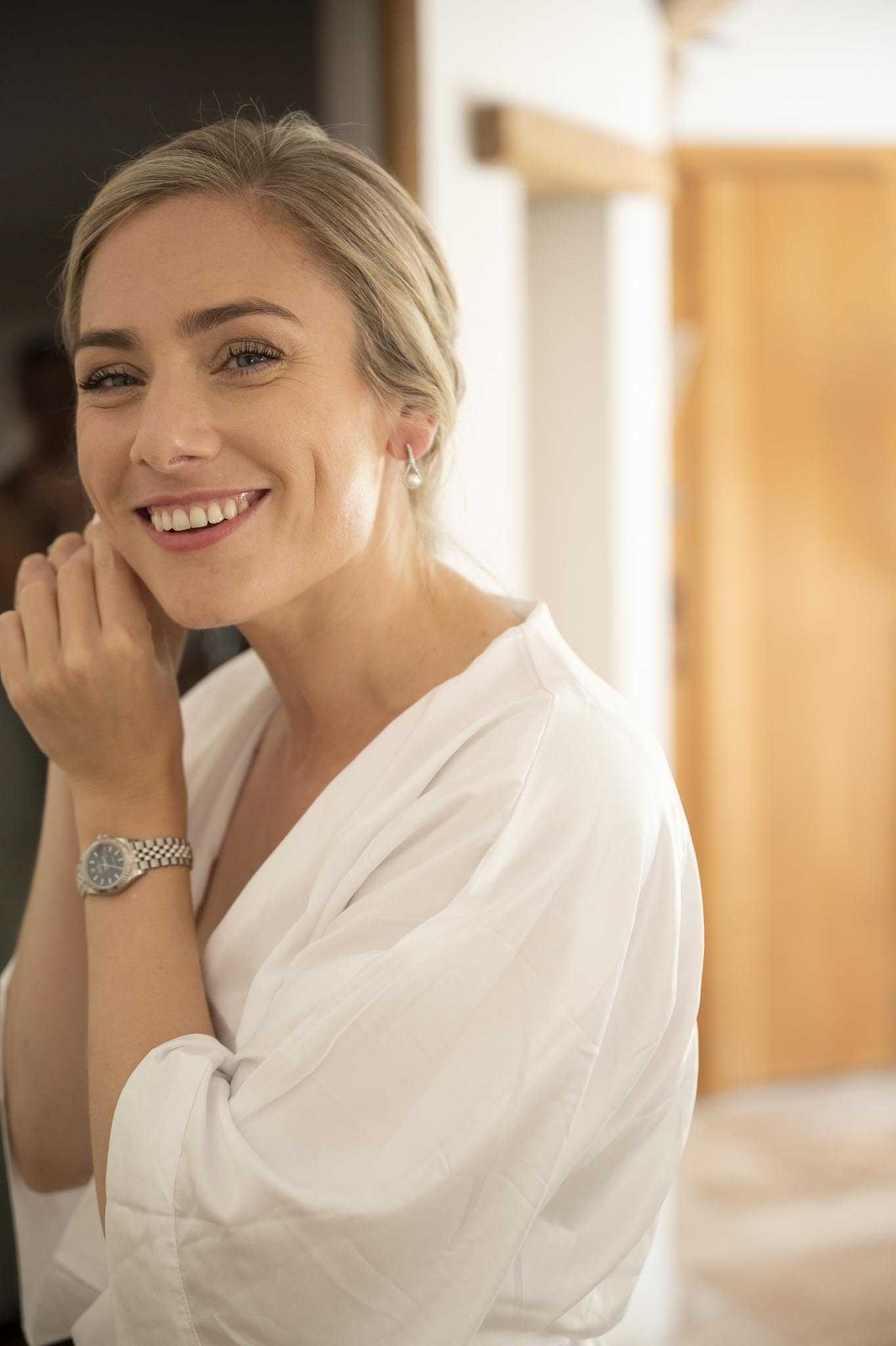 A close-up portrait of a bride during the getting-ready phase, wearing a white wrap robe with her hair pulled back in a low updo. She is smiling and has her hand raised near her face, wearing a silver bracelet watch and a pearl drop earring. Her makeup is natural with a soft pink lip. The background is softly blurred, showing a warm-toned wooden door and light-filled interior space.