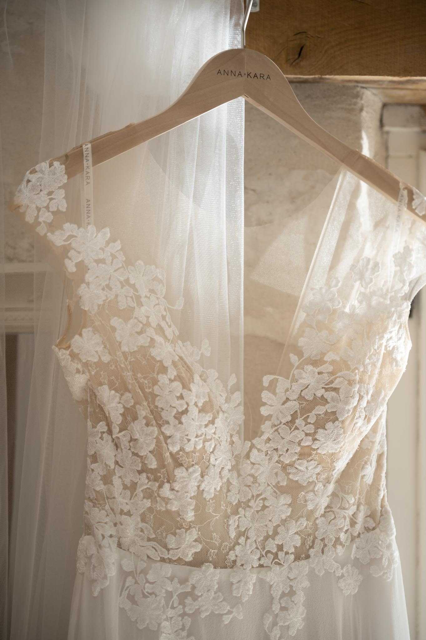 A close-up detail shot of a bridal gown hanging on a branded wooden hanger labeled 'Anna Kara,' with the same branding visible on the dress straps. The gown features ivory floral lace appliqué over a nude/champagne underlining, with cap sleeves and a deep sweetheart neckline, and a flowing tulle skirt and veil draped behind it. The dress is photographed indoors against a softly lit window, creating a warm, backlit effect that highlights the delicate lace texture. The composition is a tight portrait-orientation detail shot focusing on the bodice of the dress.