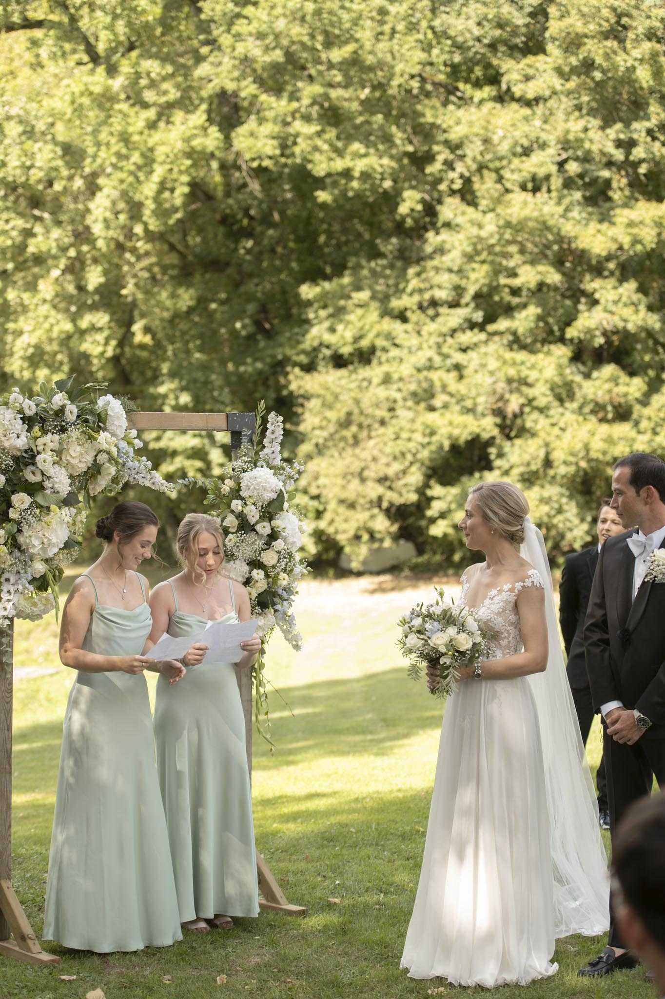 An outdoor garden ceremony is taking place on a lawn, with two bridesmaids in sage green satin slip dresses reading from a paper — likely delivering a reading or vows — while the bride listens and smiles. The bride wears a white gown with a lace cap-sleeve bodice and a flowing chiffon skirt, paired with a long veil, and holds a bouquet of white roses, ranunculus, and eucalyptus. To her right, the groom is visible in a black tuxedo with a white bow tie, accompanied by another guest in formal wear. Behind the bridesmaids stands a wooden arch decorated with white hydrangeas, white ranunculus, baby's breath, and greenery. The overall palette is white, sage, and soft green, with a classic garden wedding style. Medium wide shot capturing the ceremony party at the altar.