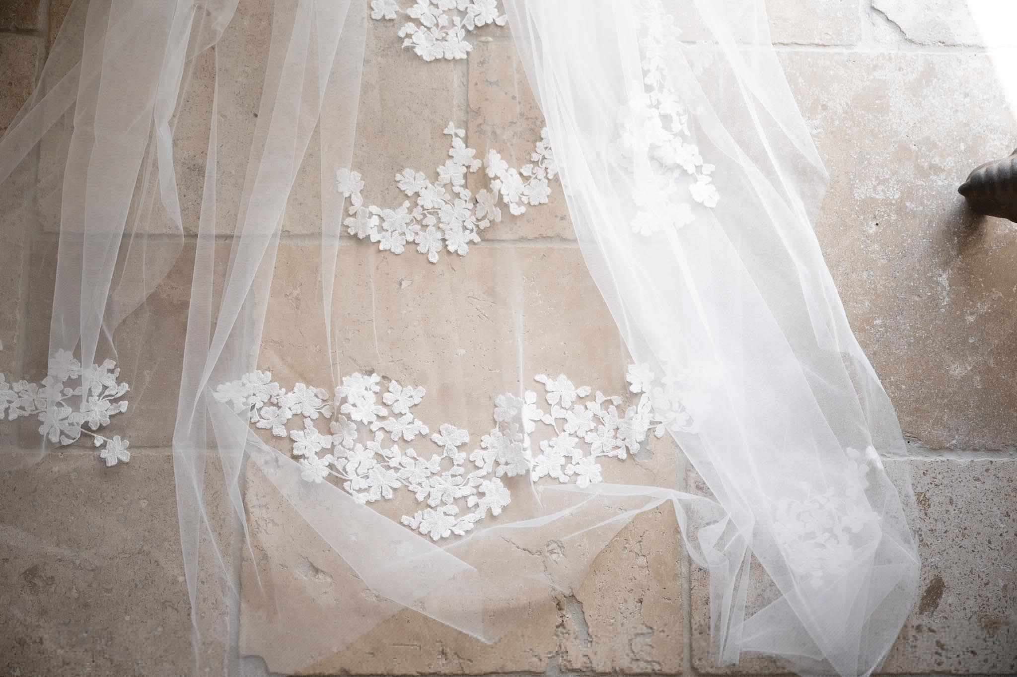 A close-up detail shot of a white bridal veil with floral lace appliqués along its edge, laid out against aged limestone or travertine flooring. The veil is made of fine ivory tulle with clusters of small white fabric flowers scattered across the border in a trailing pattern. The shot is taken from above at an angle, highlighting the delicate texture of the appliqués against the pale, worn stone surface. A small dark metal object, possibly a door fitting, is partially visible in the upper right corner.