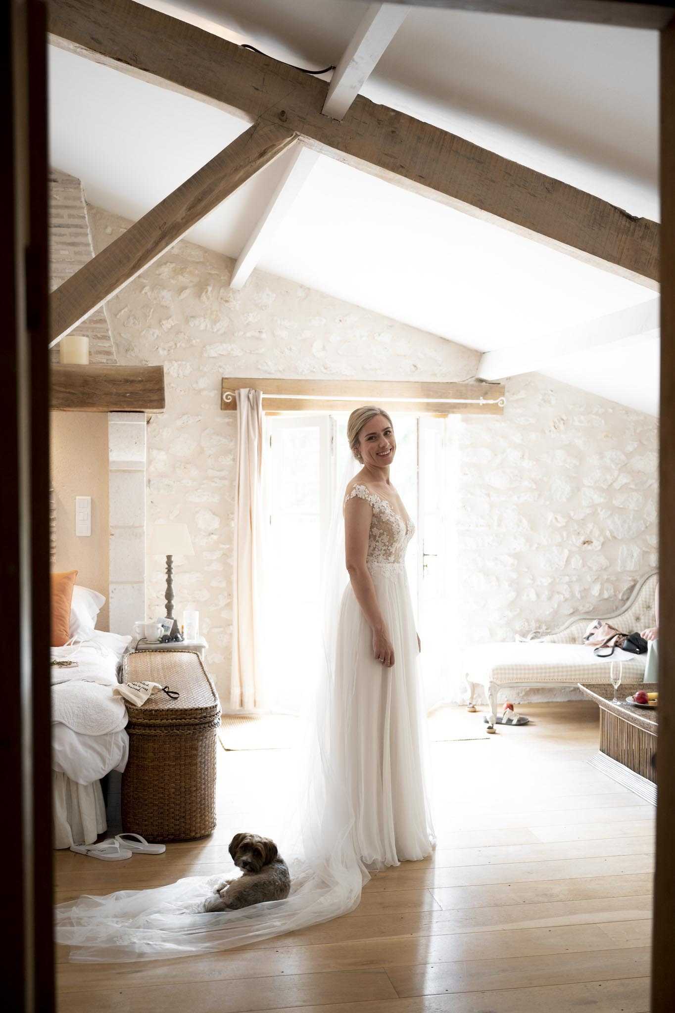 A bride stands smiling in a rustic attic-style bedroom with exposed wooden beams and stone walls, photographed through a doorframe in a medium full-length portrait shot. She is wearing a white gown with a lace illusion bodice, cap sleeves, and a flowing tulle skirt with a long train. A small brown wire-haired dog is lying on the train of her dress on the wooden floor beside her. The room is naturally lit by a bright window behind her with cream curtains, and features a wicker trunk, a bedside table with personal items, a champagne flute on a side table, and a bed with white linens and orange accent pillows, suggesting a getting-ready setting in a French countryside venue.