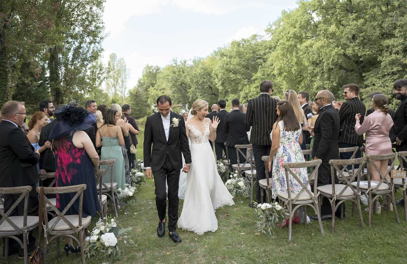 The bride and groom walk back down the aisle together following an outdoor ceremony, surrounded by approximately 30–40 standing and cheering guests. The setting is a garden or wooded grounds with a grass aisle, and the seating consists of rustic cross-back wooden chairs arranged in rows on either side. The bride wears a white A-line gown with a lace illusion bodice and a flowing skirt, her hair pinned up, and she waves to guests while holding the groom's hand. The groom is dressed in a black tuxedo with a white boutonniere. Aisle décor consists of small clusters of white peonies, white spray flowers, and eucalyptus greenery placed at the base of each row of chairs. Guests are dressed in a mix of formal attire including a bold striped blazer, a floral midi dress, a sage green dress, and a feathered navy hat. The image is a wide, eye-level shot capturing the full recessional moment with the couple centered in the frame.