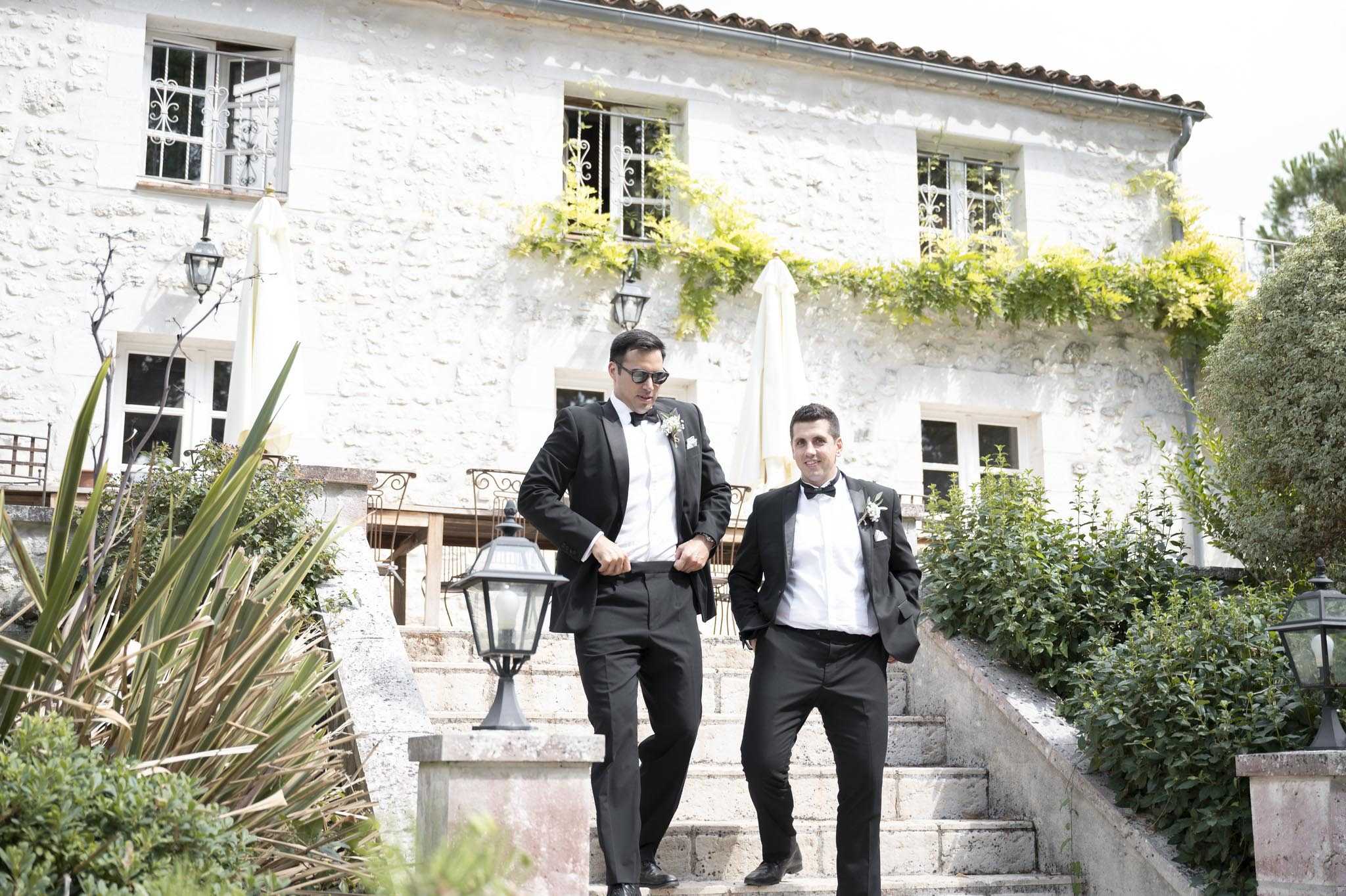 Two men dressed in matching black tuxedos with white dress shirts, black bow ties, and white floral boutonnieres are posed on the exterior stone steps of a French country manor or mas, likely the groom and a groomsman. The taller man wears dark sunglasses and adjusts his jacket cuffs while the second man stands relaxed with one hand in his pocket. The two-storey whitewashed stone building behind them features wrought-iron window grilles, terracotta roof tiles, and yellow-flowering climbing vines across the facade. Black iron lantern post fixtures line the steps, and manicured shrubs frame the staircase. The shot is a medium-wide outdoor portrait taken in natural daylight.